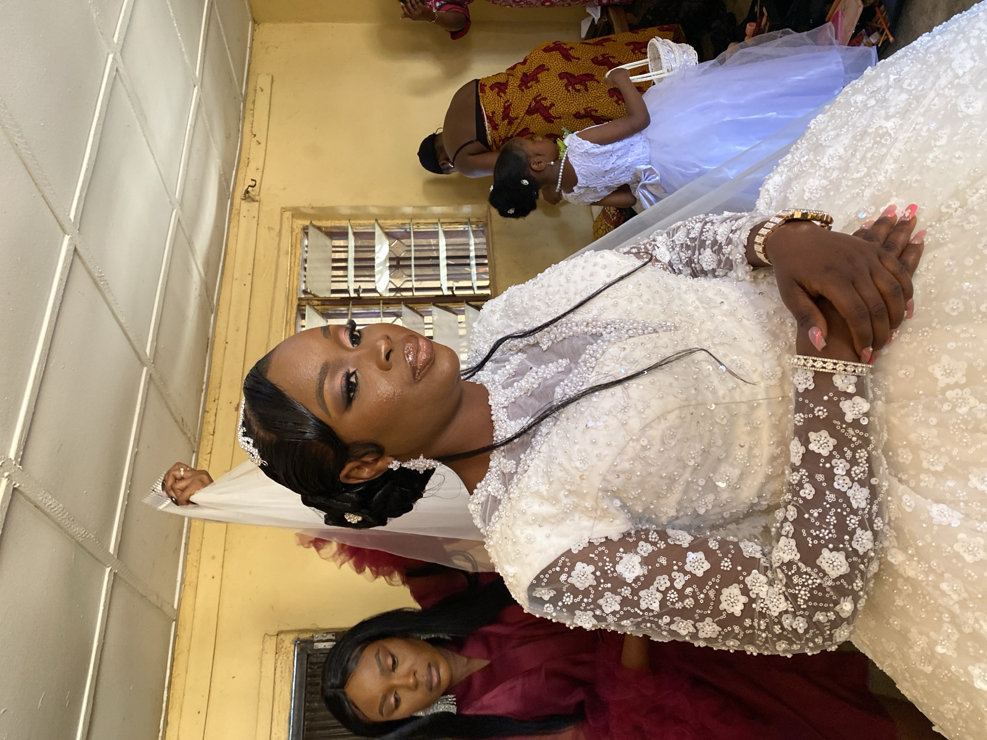 The image depicts a woman, likely a bride, in a white, heavily embellished wedding gown with lace sleeves. She is wearing a headpiece and ornate bracelets. Her nails are manicured. There is a woman in a maroon dress below her and a flower girl wearing a white dress and pearl necklace being attended to in the background. The setting appears to be indoors, possibly in a dressing area or preparatory space for a wedding. A barred window is visible in the background. The image was taken in Vom, Nigeria.