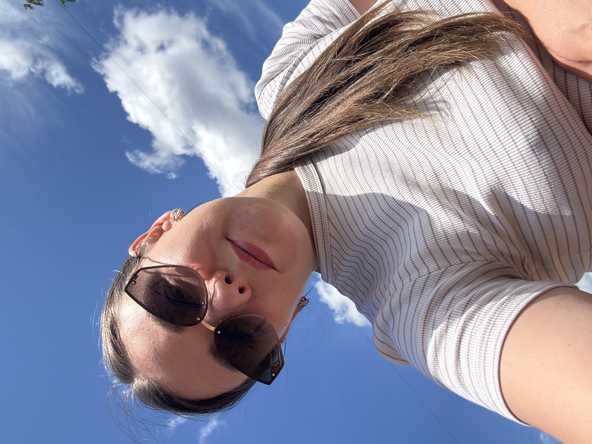 A young woman, wearing a striped t-shirt and sunglasses, is captured in a low-angle selfie against a bright blue sky dotted with white clouds. Her hair is pulled back into a ponytail. The setting appears to be outdoors, possibly in Chaikovsky, Russia, given the bright daylight and cloudy sky. The image is a close-up shot, focusing on the woman's face and upper body.