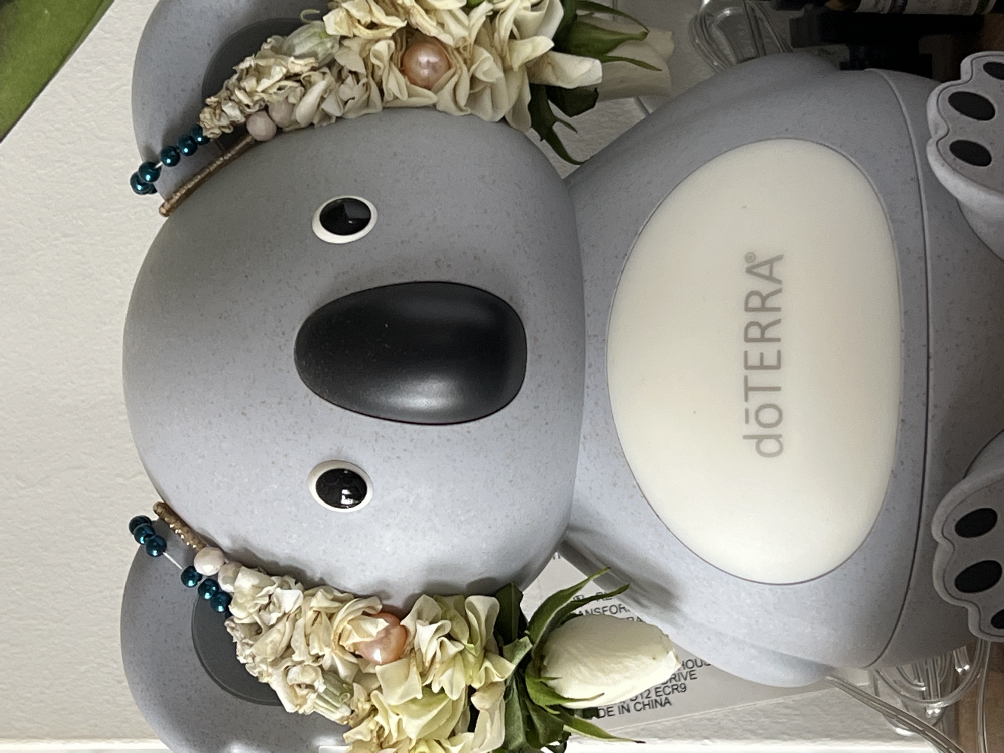 The image shows a close-up, frontal view of a grey koala-shaped diffuser. The diffuser has a large, oval-shaped white panel on its belly with the logo "dōTERRA" printed in grey text. The koala has two large, dark eyes and a prominent black nose.
Decorating the koala's ears are two floral arrangements. Each arrangement consists of dried, pale yellow and cream-colored flowers, small pearl-like beads, and some green leaves. One of the floral arrangements on the left ear has a small, unopened white rose bud at the bottom, with text printed in black on a white sticker in the background. The text appears to be a product label, with some words partially visible, including "MADE IN CHINA."
The koala diffuser is positioned on a surface that appears to be a shelf or table. To the right of the diffuser, parts of other objects are visible, including a glass bottle with a dark liquid and a thin wire, possibly part of a string of lights. The background is a plain, light-colored wall.
There are no people or animals present in the image. The image does not provide any information about the city of Bunbeg, Ireland, or its location within it.