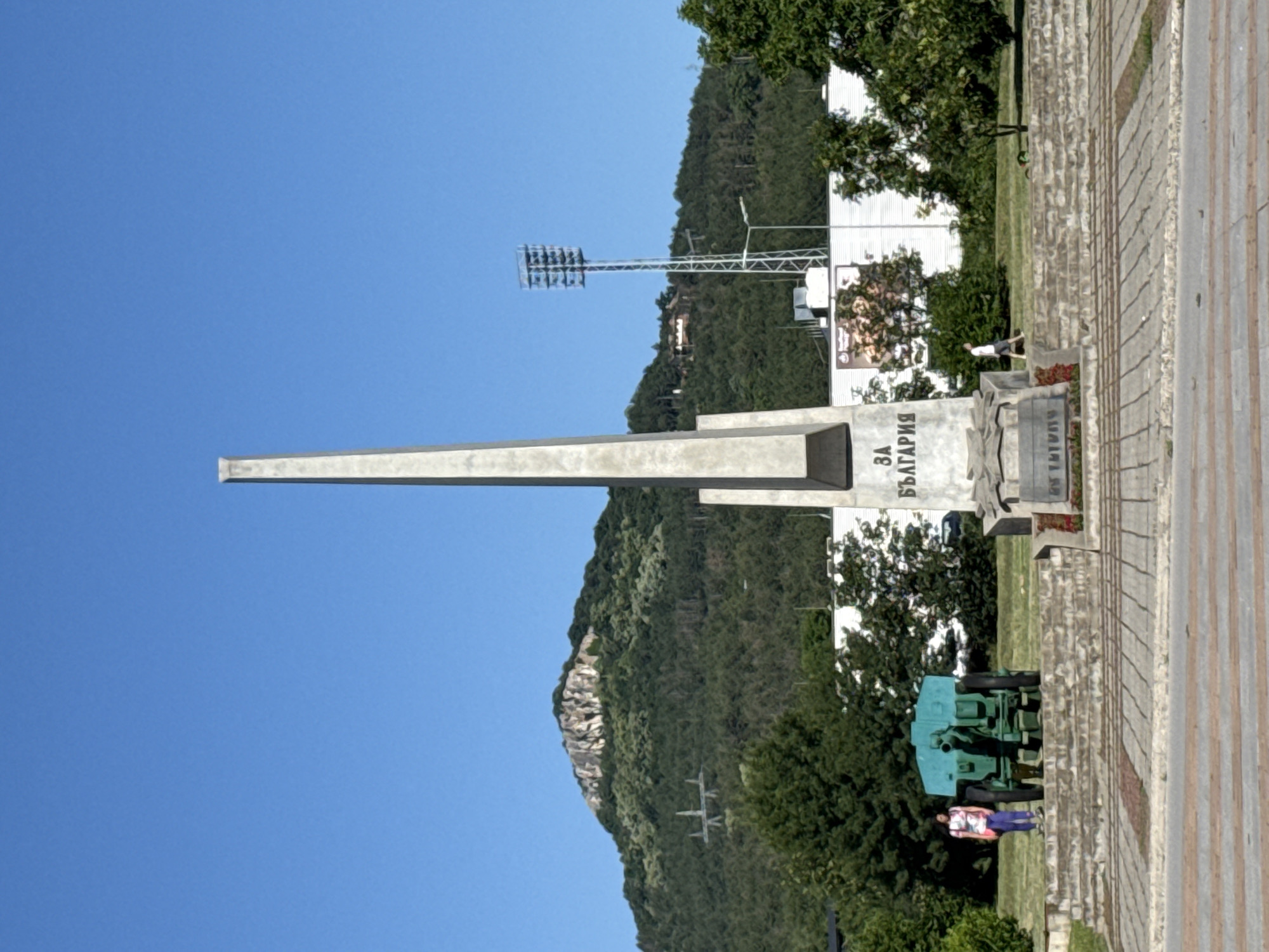 An outdoor, eye-level, vertical shot captures a monument in a park-like setting. The monument, made of light-colored stone, is a tall obelisk with a wide base. Inscriptions are visible on its front face. To the left of the monument is a bright teal-colored artillery piece with black wheels. A young woman with dark hair, wearing a white and pink patterned t-shirt and purple pants, stands to the left of the artillery piece. She is looking towards the monument. In the background, a lush green, tree-covered hill rises, with a rocky peak at its summit. A set of stadium lights is visible on the right side of the hill. A power line tower with a distinctive lattice structure is also present on the hillside. The area in front of the monument features a paved walkway and a stepped stone platform with some greenery. A person in a white t-shirt and shorts walks to the right in the middle ground. The sky is a clear, bright blue.