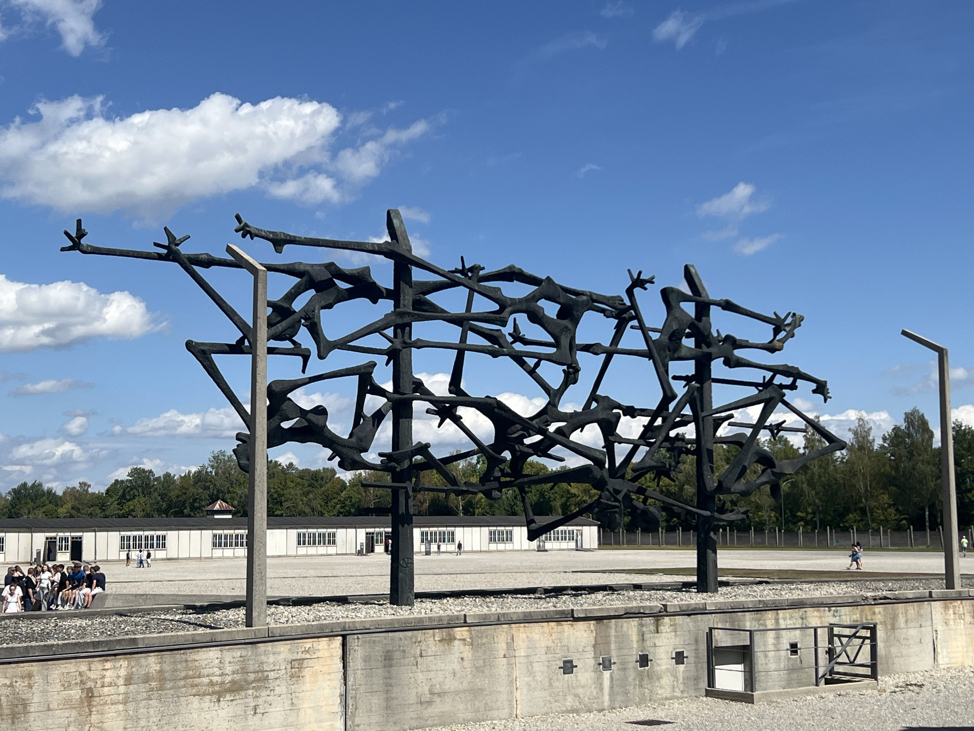 The image depicts the Memorial of the Dachau Concentration Camp in Dachau, Germany. In the foreground, a large, abstract black sculpture, consisting of intertwined human-like figures, stands prominently. This sculpture is situated in front of a concrete wall with gravel on the ground. Behind the sculpture, a vast, open, gravel-covered area stretches towards several identical, long, white barracks with dark-paned windows, characteristic of former camp structures. A line of trees forms the background, with a clear blue sky dotted with white clouds above. On the left side of the image, a group of visitors, appearing to be students, are seated on a low concrete barrier, observing the memorial. Other individuals are sparsely scattered across the open area, walking or standing. The overall scene conveys a sense of solemnity and remembrance within a historical site.