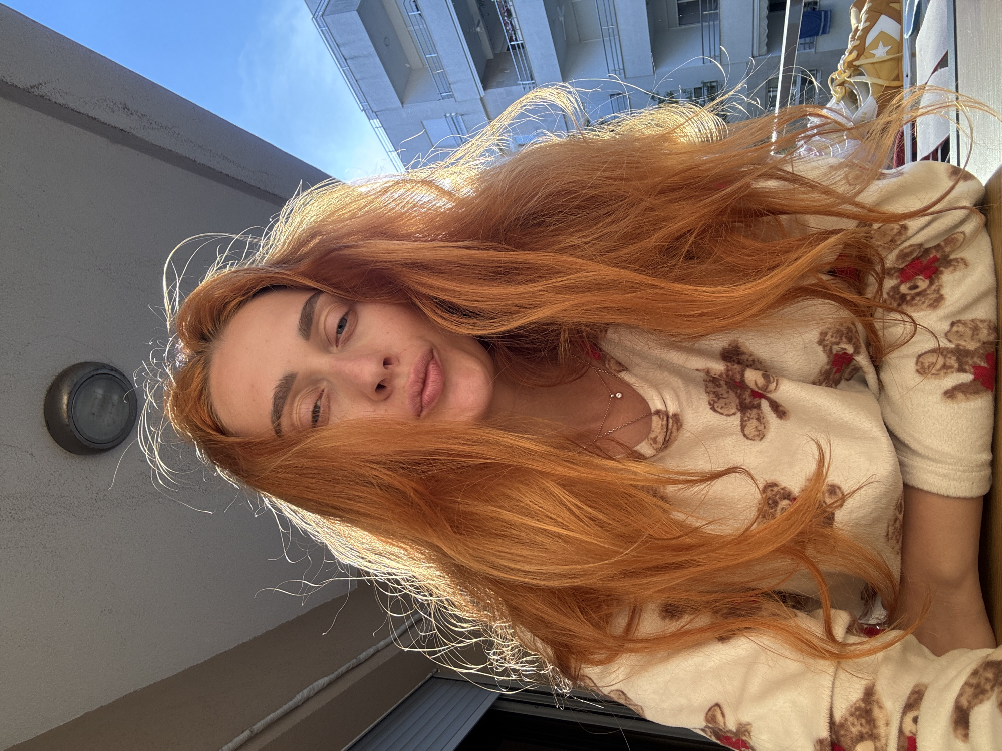 A woman with long, auburn hair is wearing a teddy bear-patterned pajama top. She is on a balcony or rooftop in Budva, Montenegro, with a building and the sky visible in the background.
