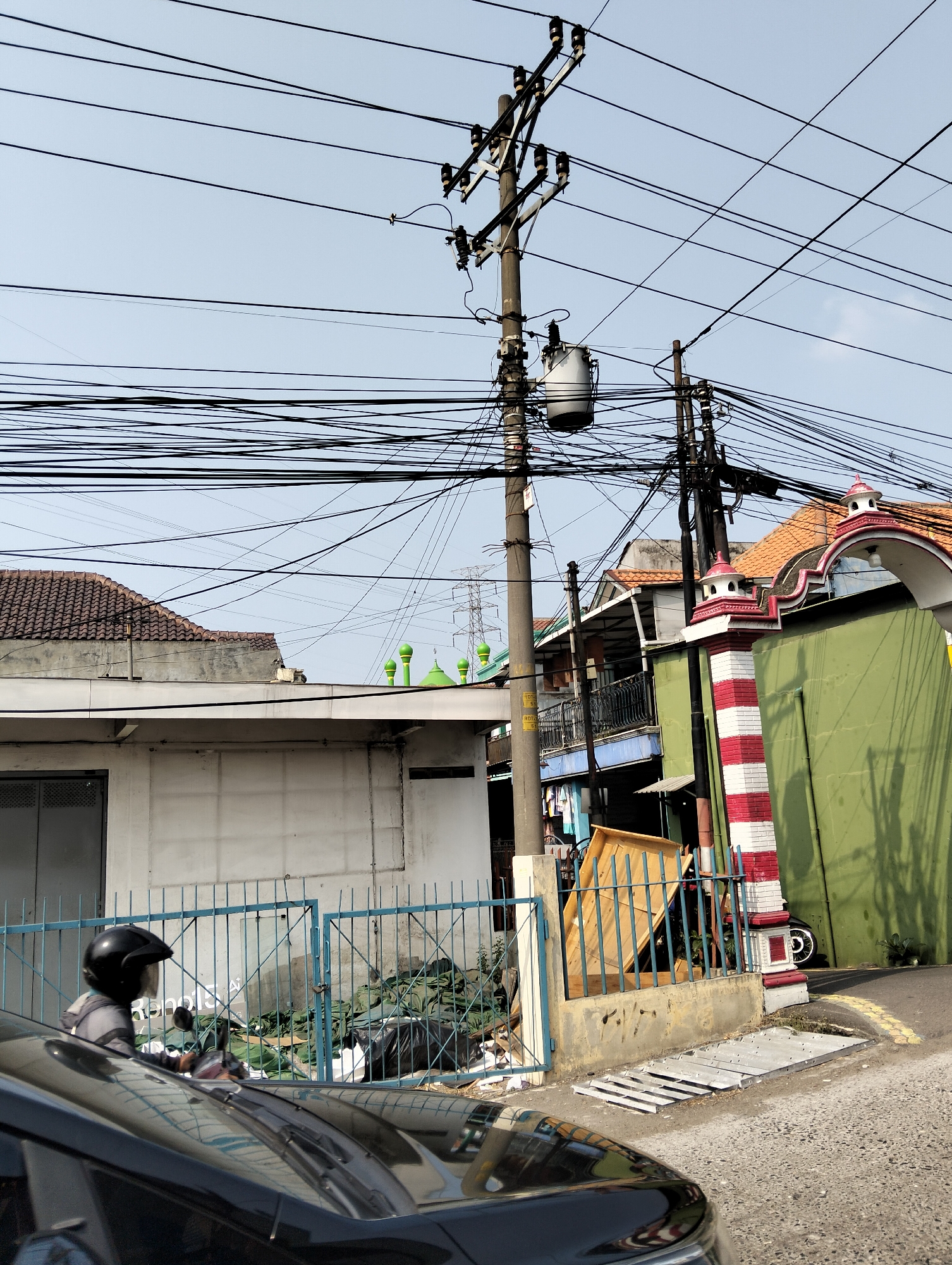 The image depicts a street scene in Semarang, Indonesia, characterized by a dense network of overhead electrical wires and a prominent utility pole. The pole supports a transformer and numerous tangled cables. In the foreground, the hood of a black car is visible, and a person on a motorcycle wearing a helmet is captured in profile. Behind them, a blue metal fence encloses an area with discarded materials, including green tarps and black trash bags. Adjacent to the fence is a green building with a red and white striped decorative pillar. Further back, several houses with tiled roofs are visible, some with balconies. Two green dome structures, likely part of a mosque, are also present in the background. The sky is clear and bright, suggesting daylight. The overall impression is one of a busy urban environment with a functional but somewhat cluttered infrastructure.
