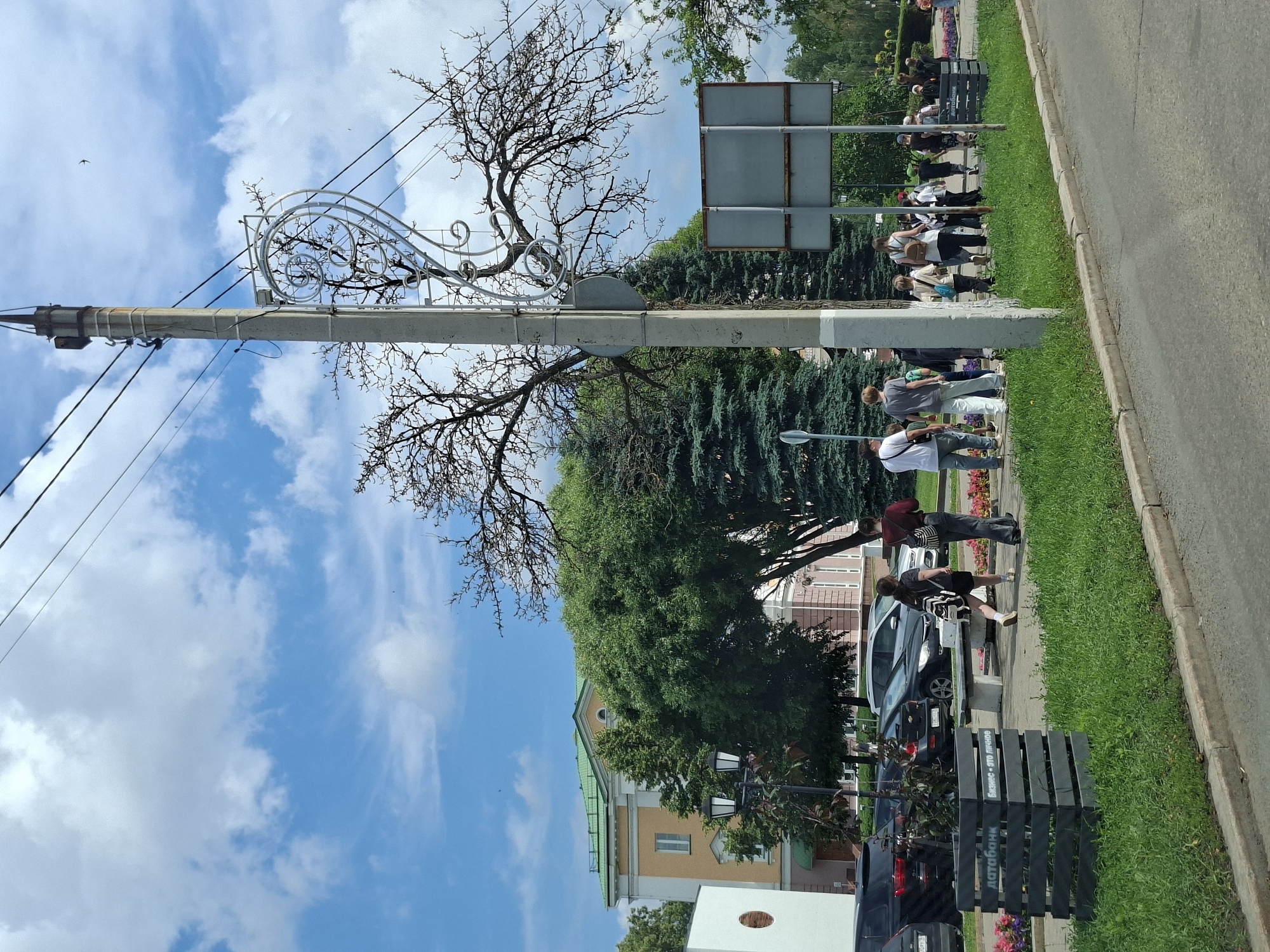 The image depicts a scene on a city street in Izhevsk, Russia, on a cloudy day. In the foreground, a paved road runs alongside a grassy area with a concrete curb. A decorative planter box with the inscription "Датабанк Бизнес — это личное" (Databank Business is personal) is visible on the grass.
A group of people are walking along the sidewalk. They appear to be of various ages and are dressed in casual clothing. Some are walking individually, while others are in pairs or small groups. Behind the sidewalk, cars are parked along the street.
In the background, a building with a yellow facade and a green roof is visible, partially obscured by trees. A lamppost stands near the building. A concrete utility pole with electrical wires runs through the center of the image, adorned with a decorative metal element shaped like a heart or swirl. Bare branches of a large tree extend across the upper right portion of the frame, suggesting it might be early spring or late autumn. Two large, gray rectangular signs are mounted on poles on the right side of the image. The sky is a mix of blue sky and scattered clouds.