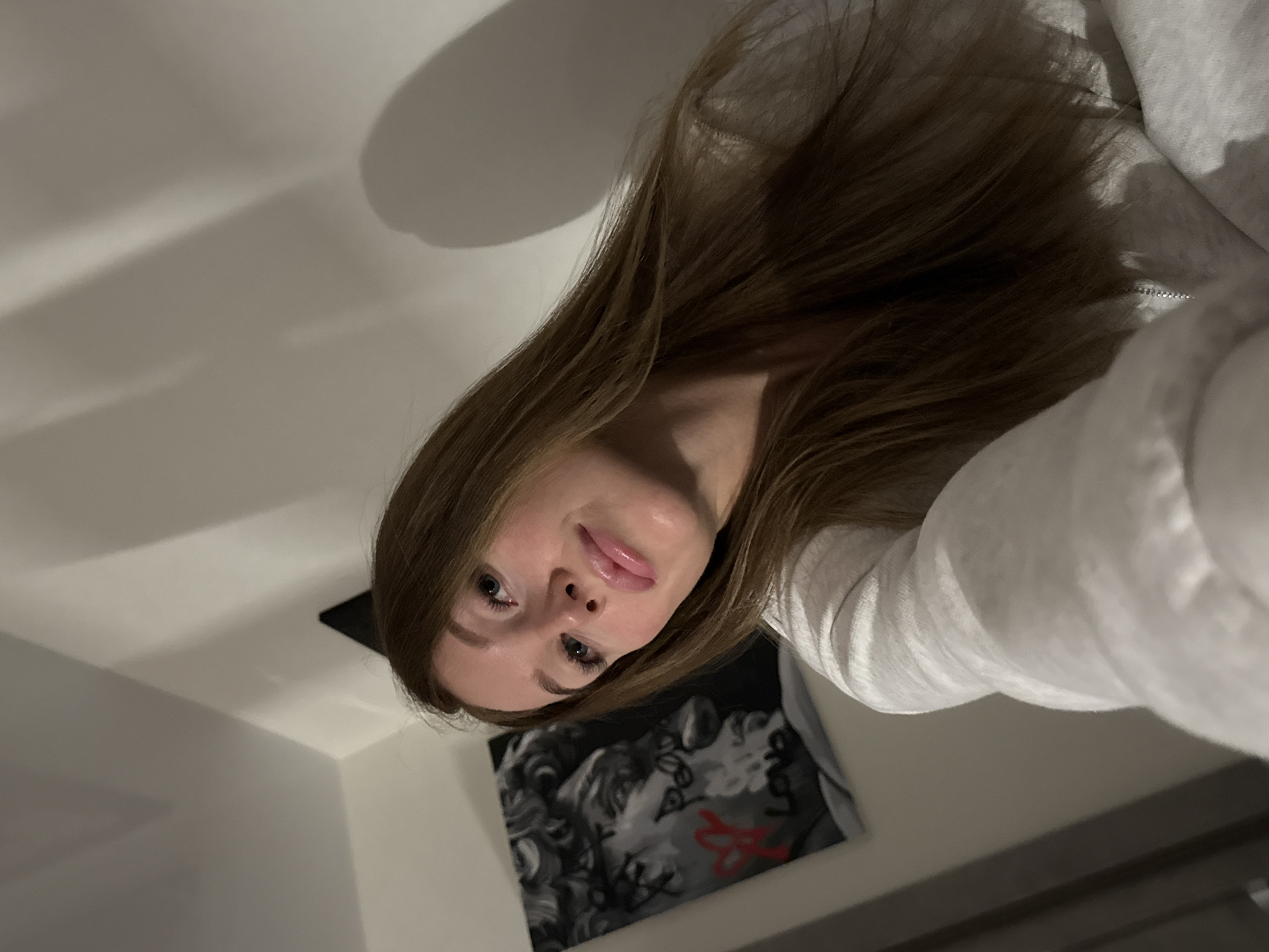 A selfie taken from a low angle features a young woman with long brown hair, fair skin, and blue eyes. She is wearing a light gray hooded sweatshirt. Behind her is a white wall with a black and white picture of a face, possibly a classical sculpture, defaced with red graffiti in the shape of a heart and the word "Love" written in an unknown language. The photo was likely taken indoors under artificial lighting. The location in the city of Chaikovsky, Russia, is not evident from the visual information.