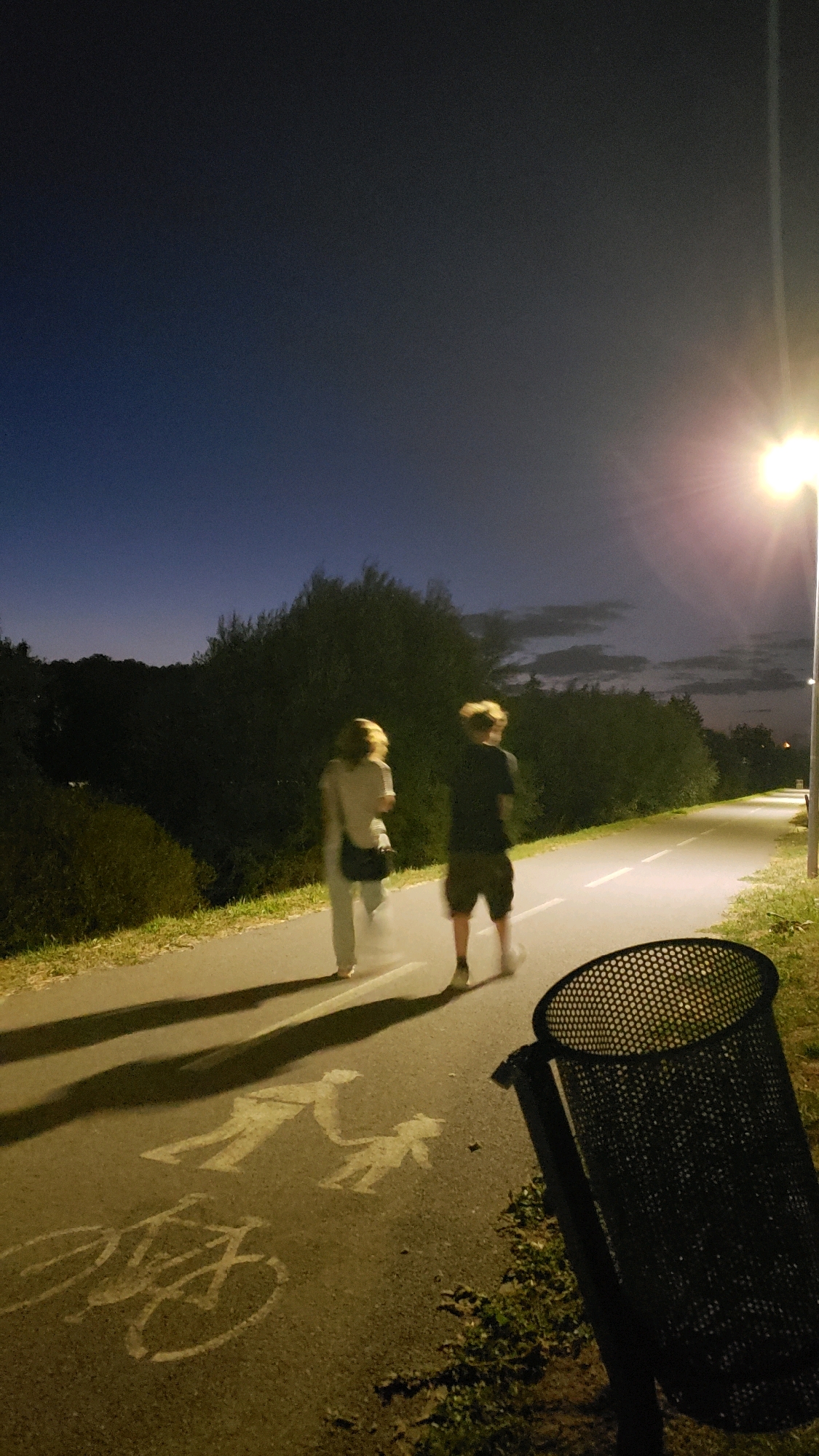 The image captures a nighttime scene on a paved pathway, likely in Prešov, Slovakia, given the provided context. Two individuals are walking away from the camera on the path. The person on the left appears to be wearing light-colored pants and a top, with a bag slung over their shoulder. The person on the right is wearing shorts and a t-shirt, with their back to the viewer. Both individuals are blurred due to motion. The pathway is illuminated by a streetlamp on the right side of the frame, which is casting a bright light and long shadows. Painted markings on the pathway include a bicycle symbol and a symbol depicting an adult with a child. The pathway is bordered by grassy areas and dense foliage. A perforated metal trash can is visible in the foreground on the right. The sky is dark with a gradient of blue and gray, suggesting twilight or late evening.