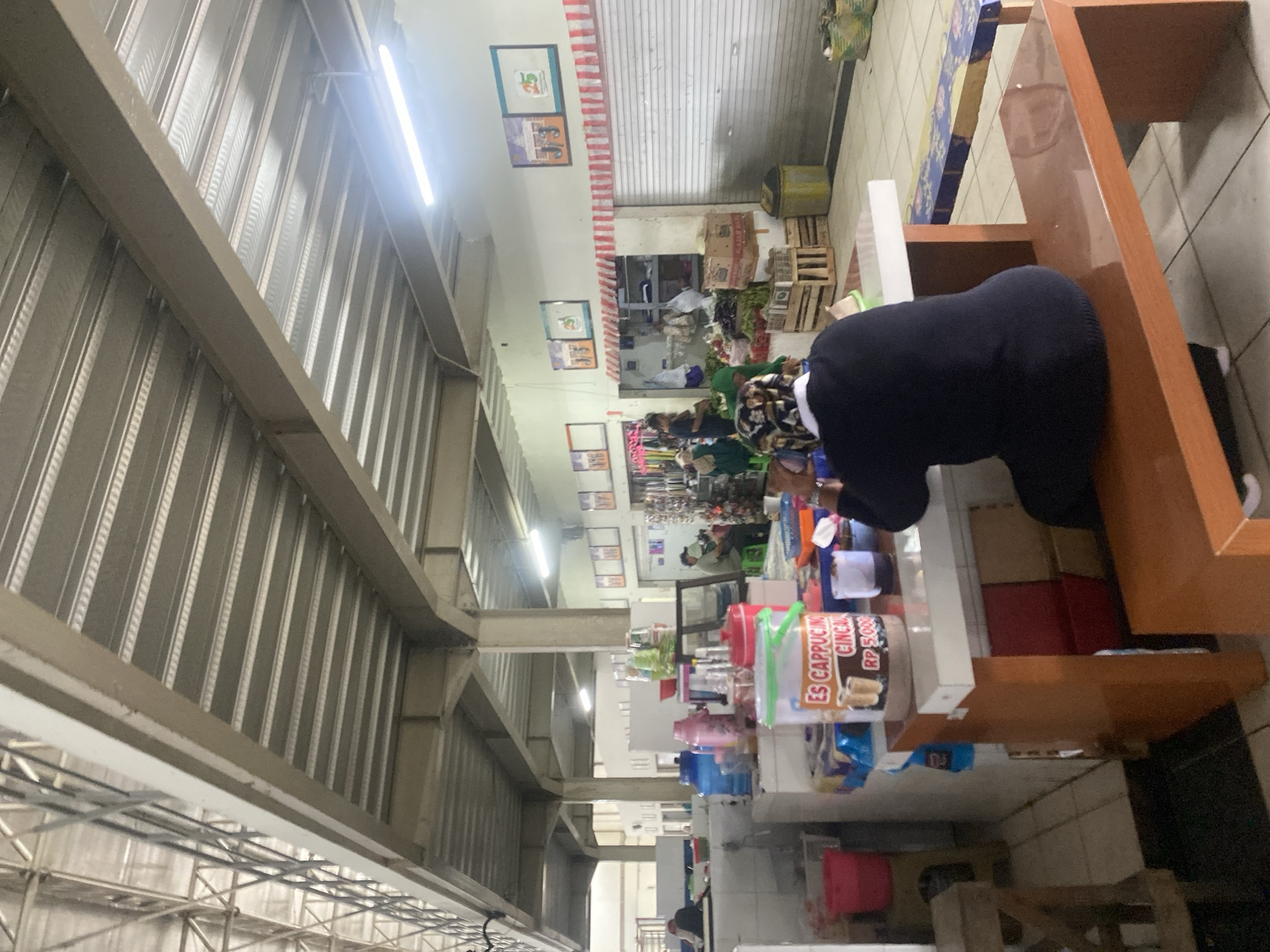The image shows a covered market or food court in Banjarbaru, Indonesia. A woman wearing a dark blue sweater and headscarf is seated at a table with a white top and wooden base, her back to the camera. On the table, there is a large plastic container with an "Es Cappucino Cincau" label, a small cup of dark liquid, and other assorted items. The table is positioned on a tiled floor. In the background, various stalls are visible, some with closed metal shutters. There are other people in the market, including a person in a green headscarf attending to a stall with produce. The ceiling of the market is constructed with corrugated metal panels and exposed beams, illuminated by fluorescent lights. The overall environment suggests a busy marketplace.