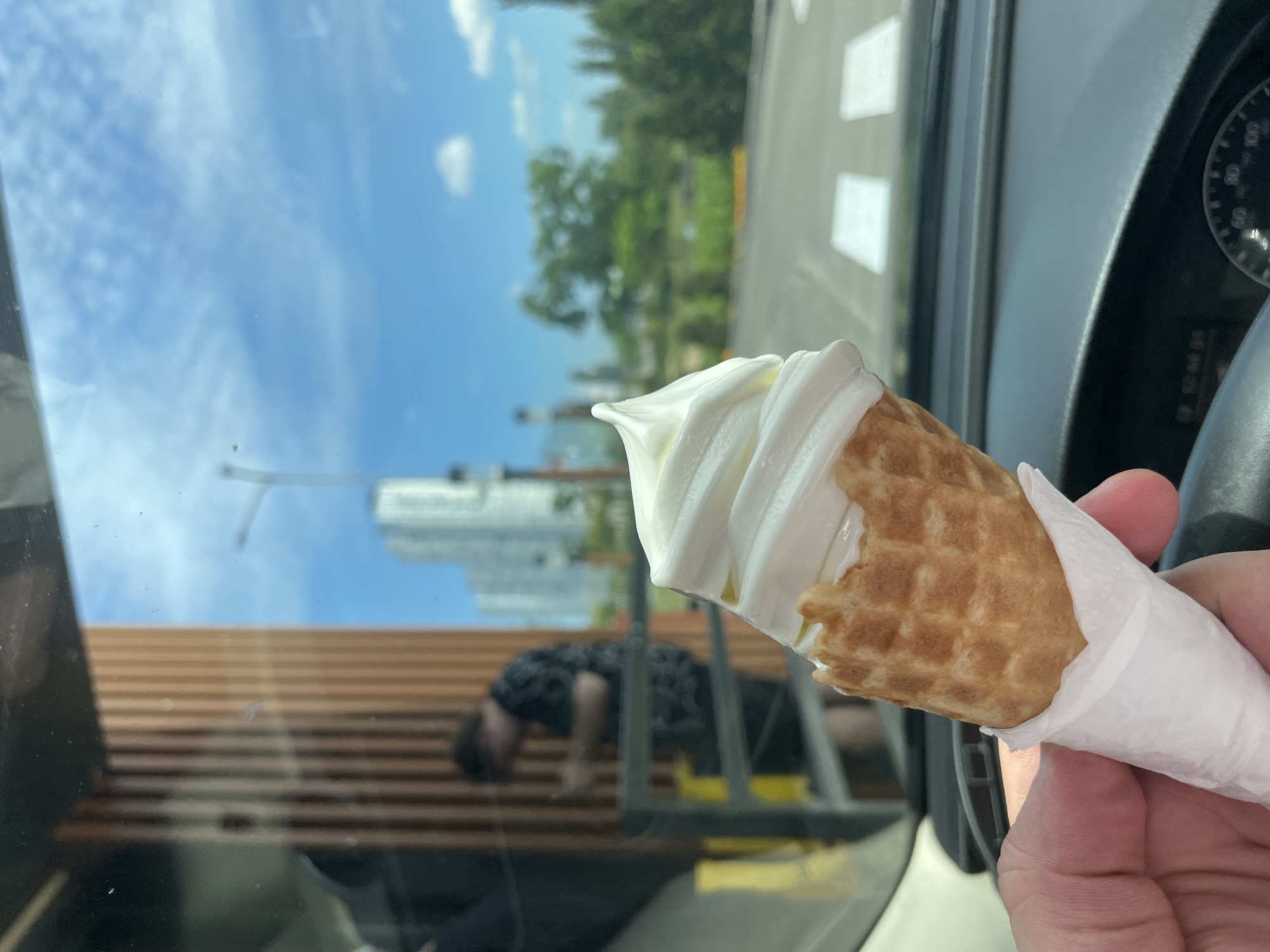 A person holds a vanilla soft-serve ice cream cone in a waffle cone, wrapped in a white paper napkin, while seated in a car. The ice cream is in focus in the foreground, obscuring the lower portion of the car's dashboard and steering wheel. In the background, seen through the car's windshield, is an outdoor setting with a modern wooden building facade on the left, a metal railing, and a person in a dark patterned shirt standing near the building. Further in the distance, there are tall buildings, trees, and a bright blue sky with scattered clouds. The scene appears to be a drive-thru or outdoor service area in Kyiv, Ukraine, evidenced by the architecture and sky conditions typical of the region.