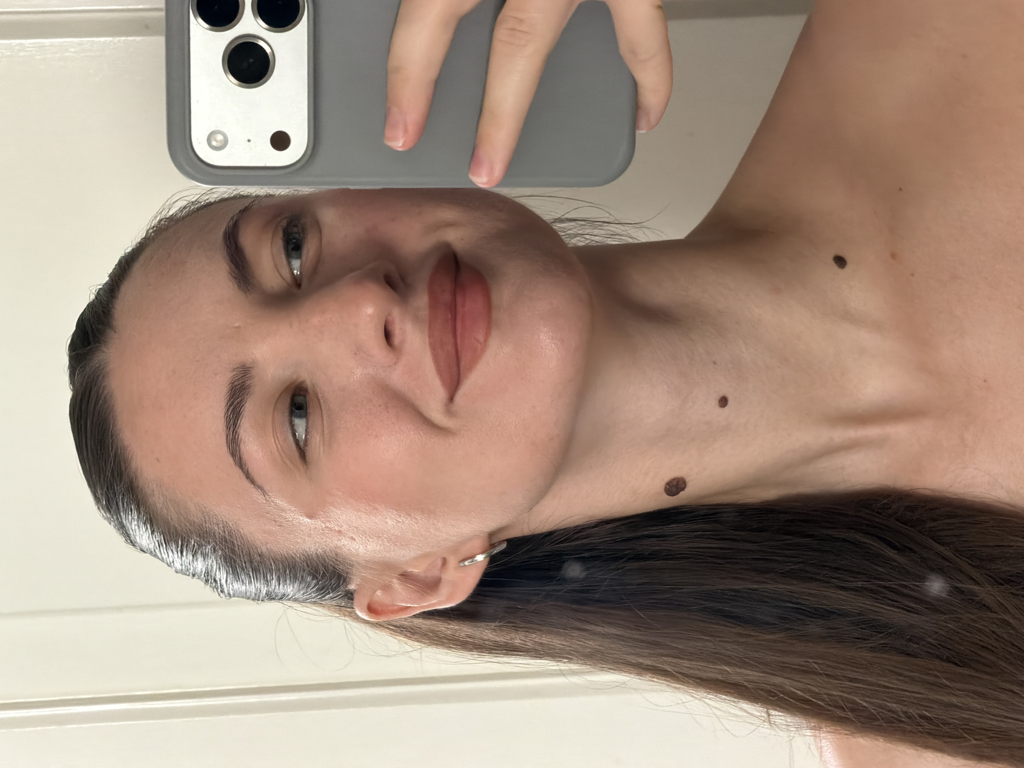 A woman is taking a selfie with her smartphone. She has long, dark brown hair pulled back into a ponytail, blue eyes, and light skin. She is wearing a simple silver hoop earring in her left ear. There are three moles visible on her neck and upper chest. The background appears to be a plain wall, possibly in a residential setting. There is no indication of the city of Sharm El Sheikh, Egypt.