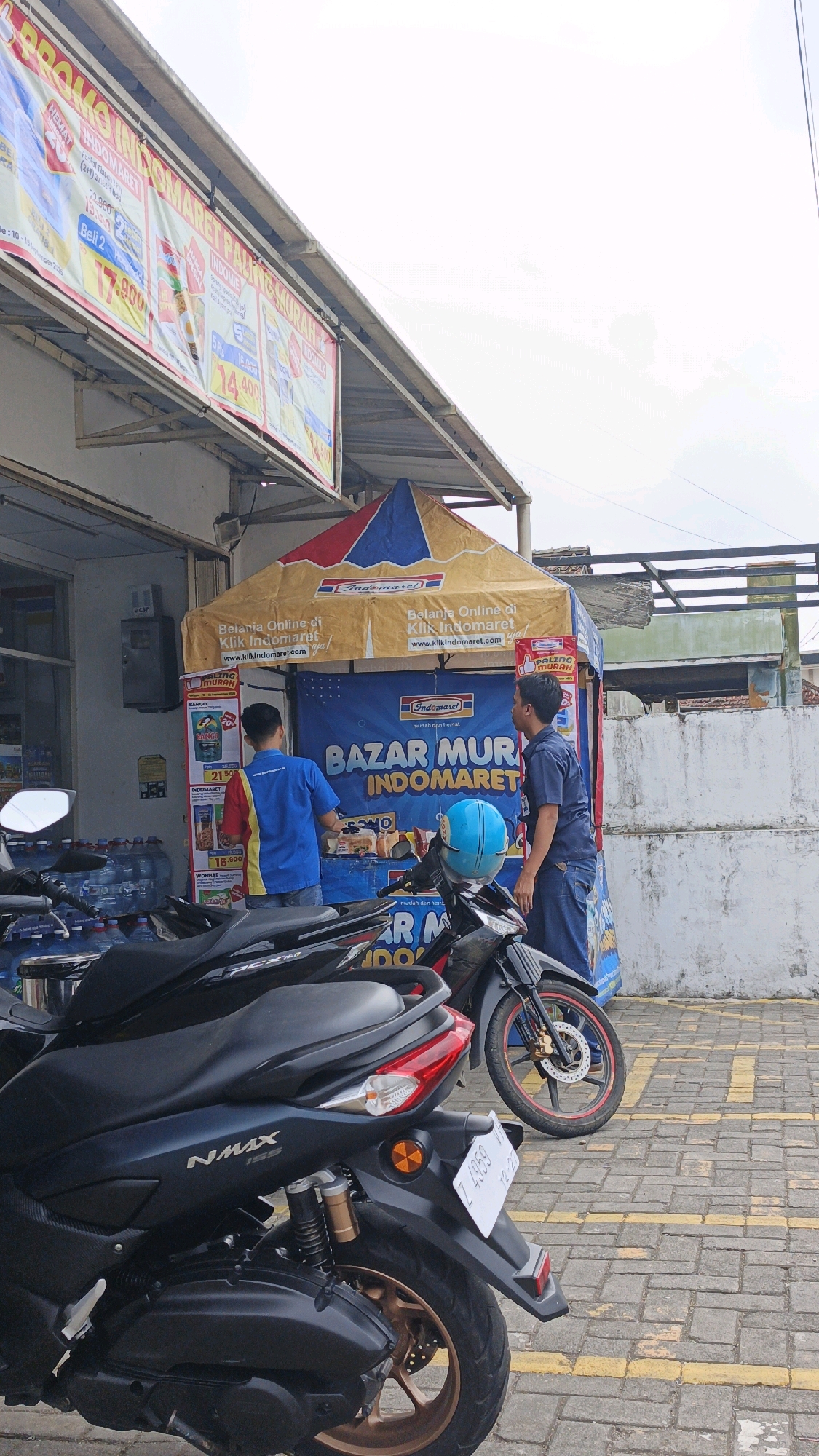 In Cibenda, Indonesia, a temporary outdoor bazaar is set up in front of an Indomaret convenience store. Two men, likely store employees, are engaged at the bazaar stall. One man, wearing a blue shirt with red and yellow accents, faces away from the camera while arranging items. The other, in a navy blue uniform shirt and jeans, stands and observes.
The bazaar tent is yellow with red and blue trim, featuring Indomaret branding and advertising for "Belanja Online di Klik Indomaret" (Online Shopping at Klik Indomaret). A blue banner behind the stall reads "BAZAR MURAH INDOMARET" (Indomaret Cheap Bazaar). Several products are displayed on shelves at the stall.
In the foreground, two motorcycles are parked. The closest is a dark-colored NMAX 155 scooter, its rear fender and license plate (Z 4959) visible. Behind it is another dark motorcycle. To the left of the bazaar, stacked bottles of water are visible through the store's window. The scene is set on a paved area with yellow painted lines, characteristic of a parking lot or pedestrian walkway. A plain white concrete wall forms the backdrop to the right. The sky is overcast.