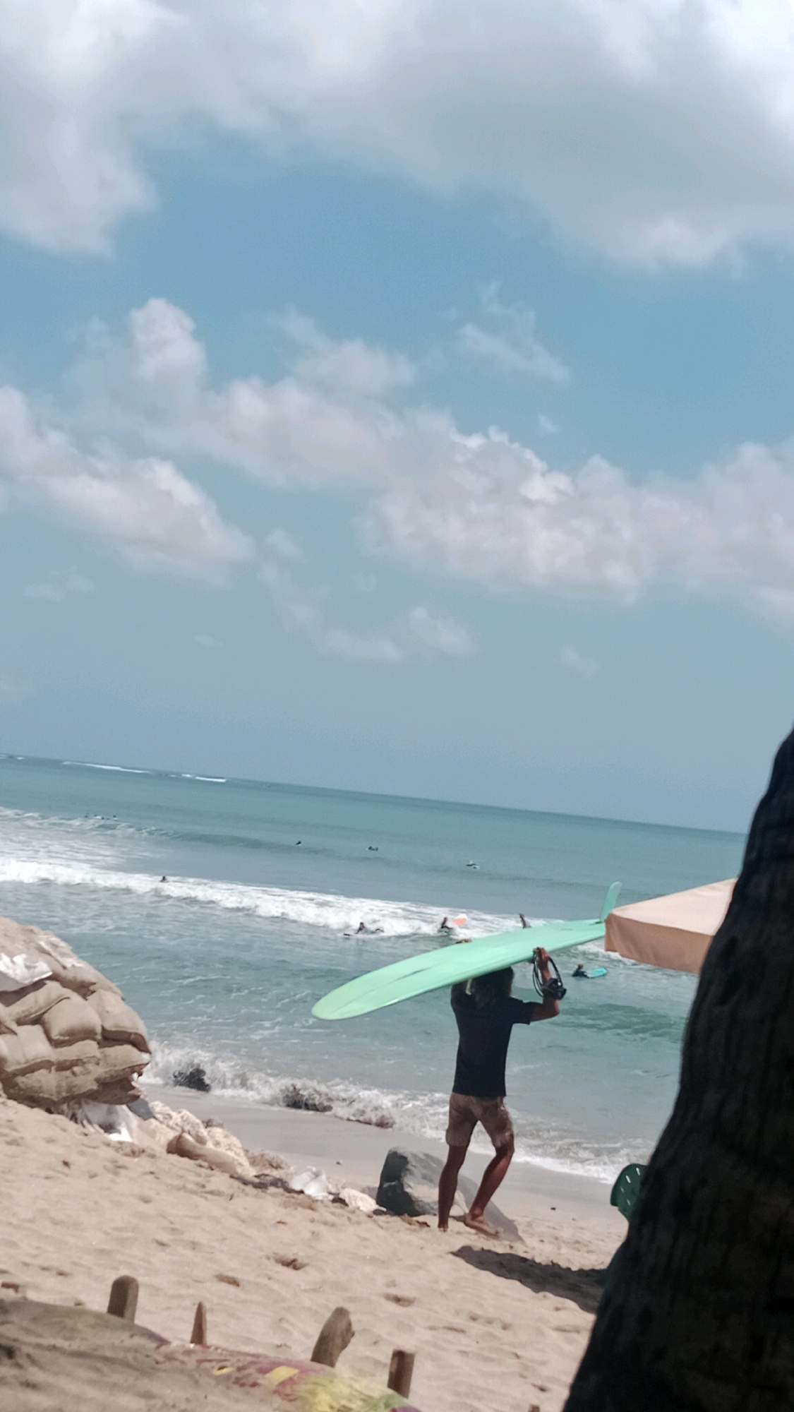 A person carries a surfboard on their head while walking on a sandy beach in Kuta, Indonesia. The ocean is visible in the background with waves breaking and several surfers in the water. The sky is blue with scattered clouds. There are sandbags and debris on the beach, suggesting a possible construction or erosion control effort. A beach umbrella and palm tree are also present. The overall scene depicts a typical day at a popular surfing beach.