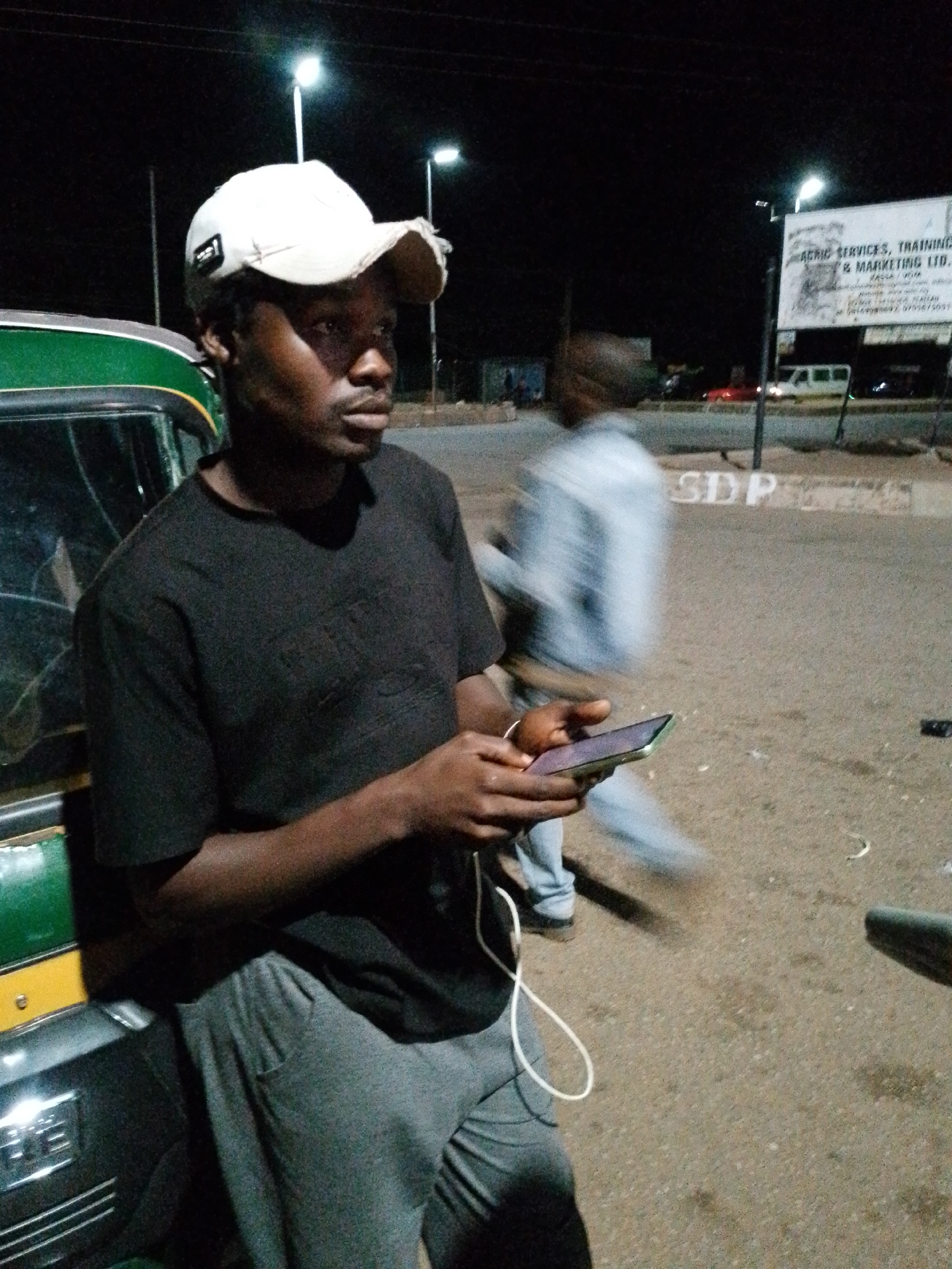 A person wearing a black t-shirt and grey sweatpants, a white baseball cap, and holding a smartphone stands next to a green and yellow vehicle. The person's left arm is bent, and their right hand is holding the smartphone. A white cord dangles from the smartphone. In the background, another person in a light-colored shirt and blue pants is blurred and walking away. A sign that reads "AGRIC SERVICES, TRAINING & MARKETING LTD." is visible in the distance, along with streetlights and a few vehicles. The ground is gravelly and appears to be a road or parking area. The scene is illuminated by artificial lighting and appears to be at night. The location is identified as Unknown, Unknown.