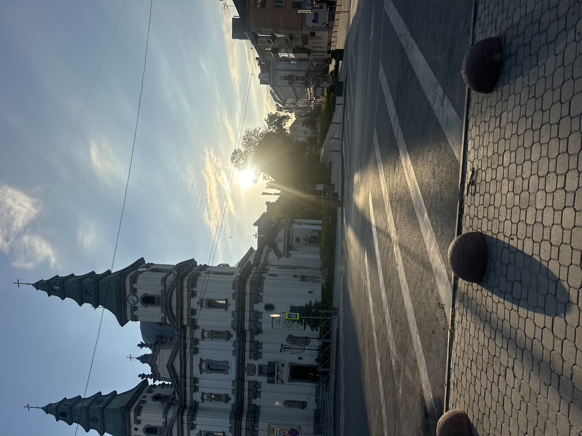 The image captures a street scene in Ternopil, Ukraine, featuring a prominent white church with two green spired towers. The sun is low in the sky, casting long shadows across the cobblestone sidewalk and the pedestrian crossing in the foreground. The church's facade is adorned with architectural details and statues. Across the street, buildings line the cityscape, with some showing signs of wear. The sky is a bright blue with wispy clouds, and power lines are visible overhead. Several round, dark bollards are placed along the edge of the sidewalk. The overall atmosphere is calm and serene, illuminated by the warm glow of the setting sun.