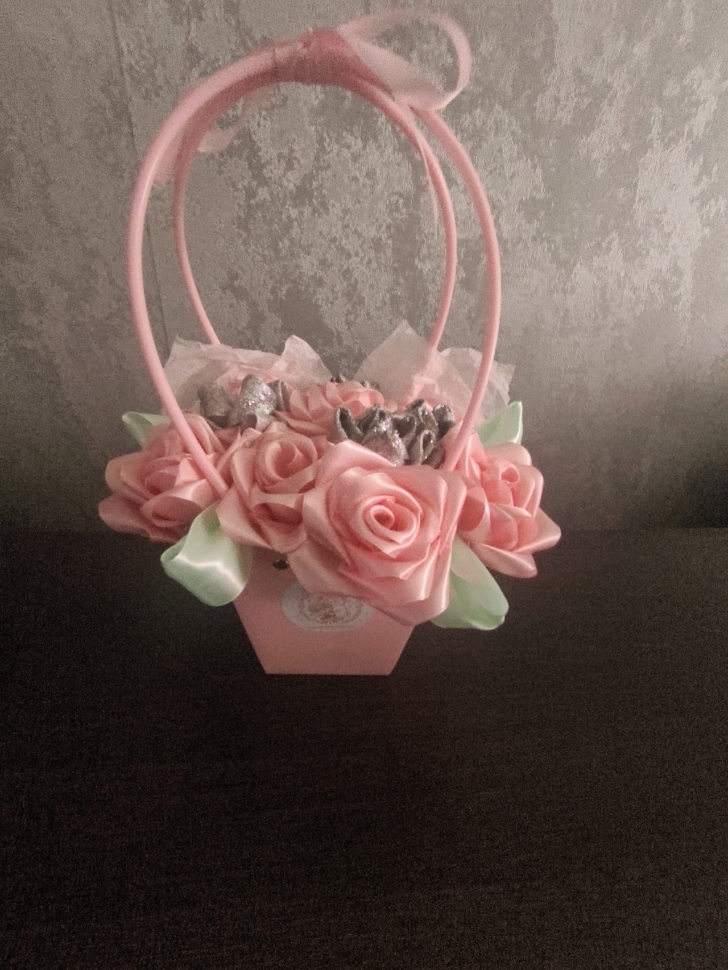 A pink artificial flower arrangement in a pink hexagonal basket with a pink handle is displayed against a textured grey wall and a dark brown surface. The flowers are predominantly pink roses made of satin, with some light green satin leaves. Silver glittery accents are interspersed among the flowers. A pink ribbon is tied to the top of the handle. The basket appears to have a decorative circular emblem on one side. The lighting suggests an indoor setting, likely a home or craft studio. There are no people or animals present. The specific location in Lisakovsk, Kazakhstan cannot be determined from the visual information provided.