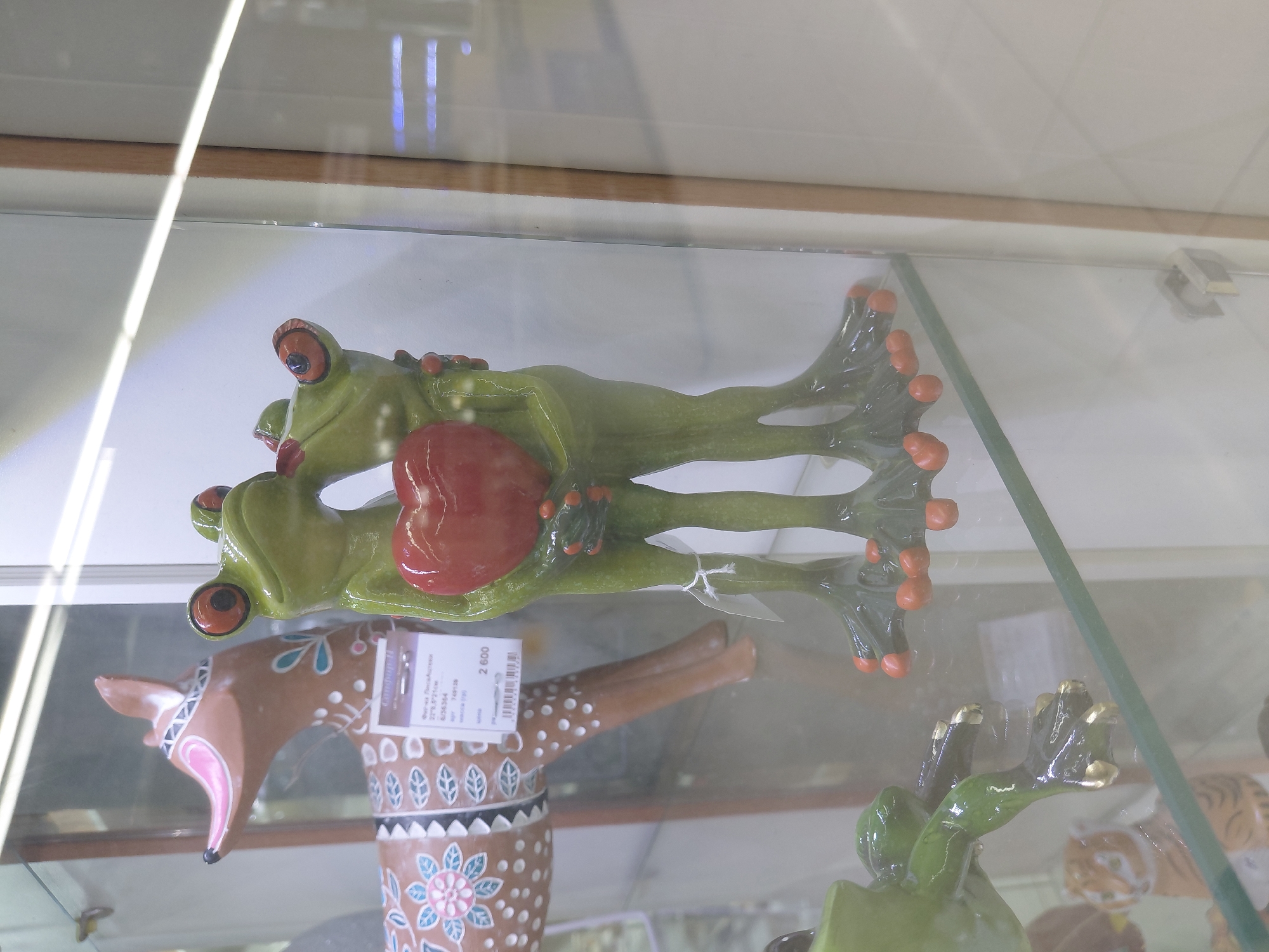 A statue of two green frogs embracing and holding a red heart is displayed in a glass cabinet. The frogs have large orange eyes and red toes. To the left of the frogs, part of a brown decorative deer statue is visible. A price tag is attached to the deer statue. The background is blurred, suggesting an indoor setting like a store.