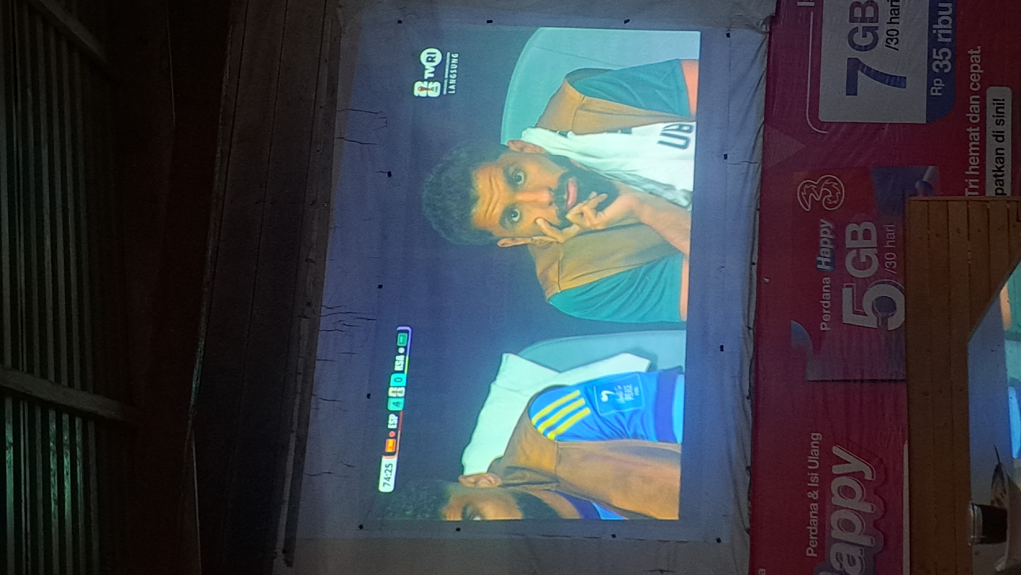 The image shows a projection screen displaying a football match. The score on the screen shows ESP 4 and KSA 0, with the time at 74:25. Two men are visible on the screen, seated. One man on the left is wearing a blue jersey with yellow stripes and a brown vest. The other man on the right is wearing a teal shirt with a brown vest and a white towel draped over his shoulder, resting his chin on his hand. A logo for TVRI LANGSUNG is visible in the top right corner of the screen. Below the projection screen, a red banner with white text advertising "Perdana & Isi Ulang Happy 3" and data packages like "5 GB /30 hari" and "7 GB /30 hari" is visible. The text "Rp 35 ribu" and "Tri hemat dan cepat. patkan di sini!" are also present on the banner. The scene appears to be outdoors, with a dark, wooden ceiling structure visible above the projection. The overall setting suggests a casual viewing of the match, possibly in a public space or an informal gathering. Based on the context provided by the banner and the commonality of such advertisements in Indonesia, the location in the city of Payagaboh, Indonesia, is inferred.