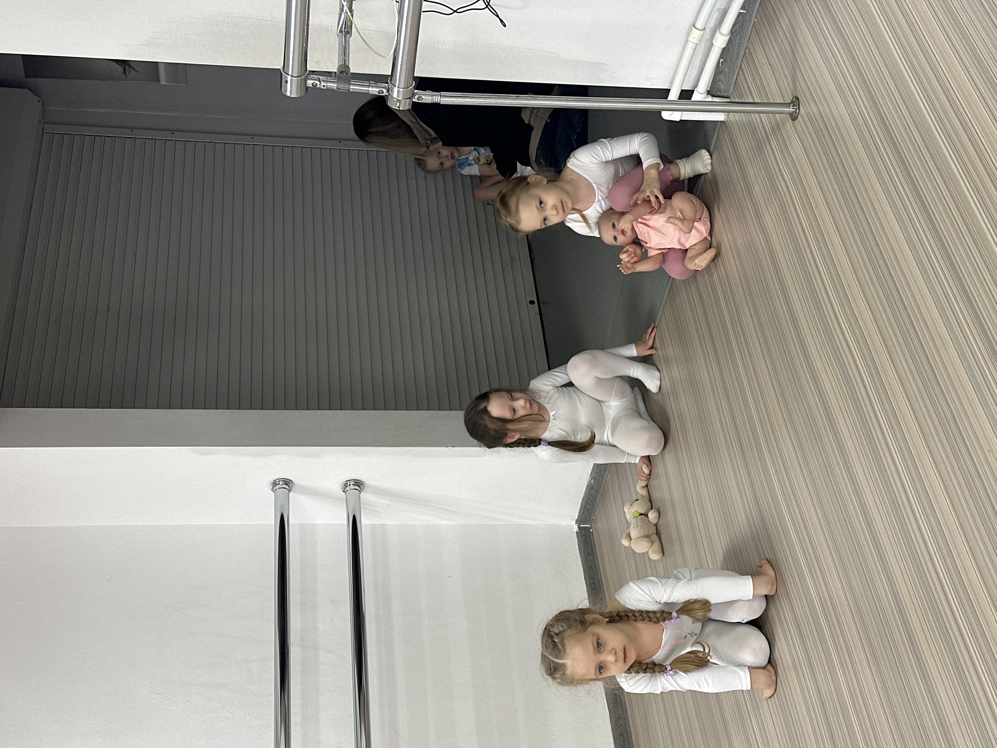 This image depicts an interior scene, likely a dance or fitness studio, in the city of Votkinsk, Russia. The foreground features three young girls, estimated to be between four and six years old, dressed in white long-sleeved leotards or athletic wear, kneeling on a light-colored wood-effect laminate floor.
From left to right:
1. A girl with blonde hair styled in two pigtail braids, secured with purple ties, faces forward, looking slightly off-camera to the left. She wears a delicate necklace with a pendant. Her feet are bare.
2. A girl with dark blonde or light brown hair in two braids faces right, looking downwards. She wears white socks. Beside her, on the floor, is a light brown stuffed animal, resembling a teddy bear.
3. A girl with blonde hair and a focused expression looks directly towards the camera. She holds a realistic baby doll, dressed in a pink romper, cradled in her lap. She is wearing pink leggings beneath her white leotard and white socks.
In the background, positioned to the right of the frame, a dark-haired adult woman is seated on a darker grey floor surface, partially obscured by a chrome barre. Another young child, with blonde hair, is seated beside the woman, looking towards the foreground.
The environment includes white painted walls. Two horizontal chrome ballet barres are mounted on the left wall. On the right side, another chrome barre assembly extends vertically from the floor. A large, grey corrugated roller shutter is visible in the background, indicating a potential entrance or storage area. The flooring transitions from the striped laminate in the foreground to a plain darker grey surface in the background.