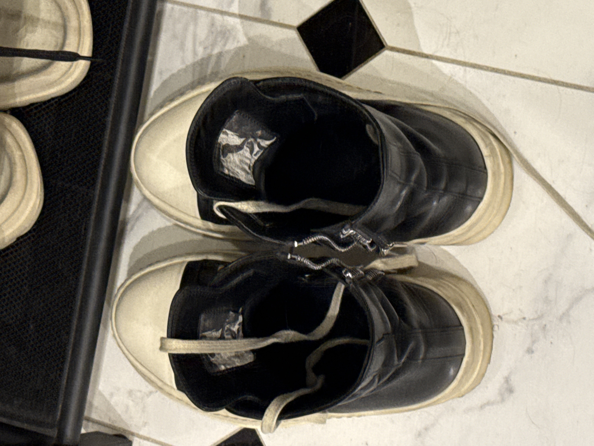 A pair of black and off-white high-top sneakers with thick soles are displayed on a tiled floor. The sneakers have zippers on the sides and laces. The floor is tiled with a pattern of white and black squares. The background shows a shelf with a textured surface, possibly a rack for shoes.