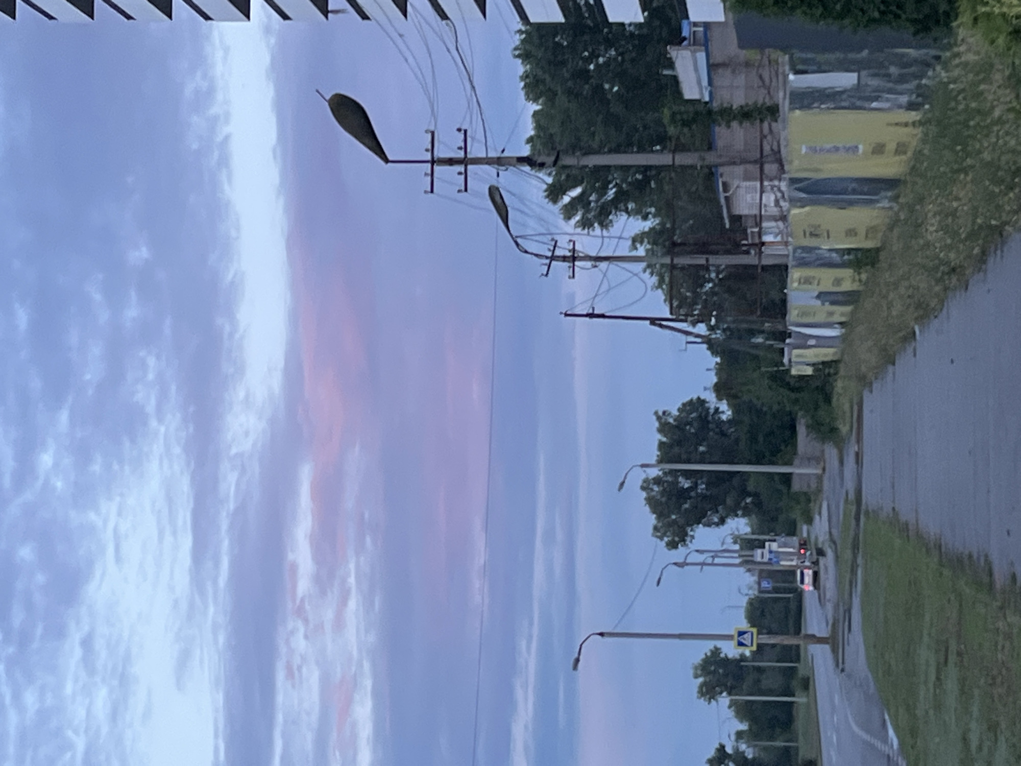 The image depicts a street scene at dusk in Dnipro, Ukraine. In the foreground, a paved sidewalk runs alongside a grassy verge, with a road to the left. Several streetlights with modern, elongated fixtures are visible along the sidewalk. A tall, modern building with a white facade and dark horizontal accents stands on the right side of the frame. Across the street, trees line the background, interspersed with some low-rise buildings. Traffic lights and road signs are present, including a pedestrian crossing sign. A white car is stopped on the road, and a truck is visible further down. The sky is overcast with shades of blue and purple, suggesting twilight. The overall impression is of a suburban or residential street in the city.