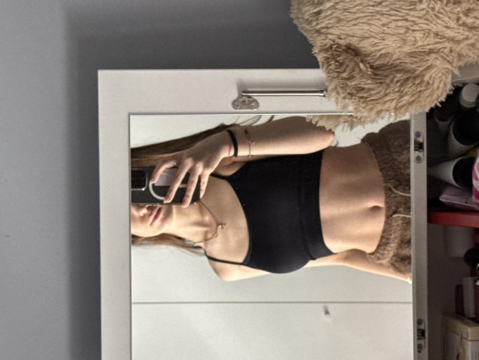 The image displays an indoor mirror selfie taken in what is stated to be Sofia, Bulgaria. A female individual is depicted in the reflection, partially obscured by a dark-colored smartphone held in her right hand, which covers her upper face from the nose upwards. She has long, dark hair. She wears a black spaghetti strap crop top, revealing her midriff, and brown, textured drawstring pants. A thin, gold-toned necklace with a small pendant is visible on her neck. Her left wrist is adorned with a black elastic band and two red string bracelets. The mirror has a white frame, and a silver-colored handle is attached to the right side of the frame, suggesting it is part of a closet or cabinet. The background reflected in the mirror consists of plain, light-colored surfaces, possibly doors or a wall. Outside the mirror's frame, a light grey wall is visible above and to the right. A large, light brown, fluffy object rests partially against the lower right exterior of the mirror frame. Below the mirror, a cluttered shelf or storage area is visible, containing various items including white boxes, a red object, and multiple cylindrical forms.