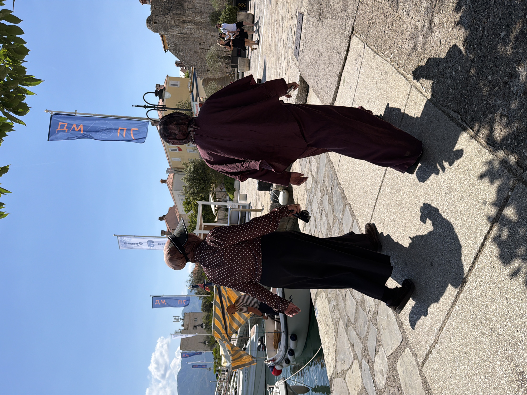 This outdoor photograph, taken in Budva, Montenegro, captures a sunny day along a paved waterfront promenade. The image appears rotated sideways but depicts a vibrant scene under a clear blue sky.
In the foreground, two individuals are walking away from the camera. The person on the left wears a loose-fitting, burgundy-colored top and trousers, and appears to be holding a small object in their right hand. Beside them, to the right, another person is seen from a side-back angle, dressed in a brown polka-dot long-sleeved shirt, a dark skirt, and a dark visor-style hat or head covering. This individual holds a small black object, likely a camera or phone, in their right hand. Both cast strong, defined shadows on the light-colored stone paving, indicating bright, direct sunlight.
Further along the promenade in the background, several other people are visible, some standing and others possibly seated. Boats are docked in the clear blue water, and buildings with traditional architecture line the waterfront. Numerous blue flags on poles are present, with one prominent flag displaying orange Cyrillic text "ДМ" (DM), a circular logo, and "ПС" (PS). Another partially visible flag has Cyrillic text "BRAVO SK". Green foliage is visible in parts of the frame, contributing to the dappled shadows on the ground. The bright lighting, clear sky, and strong shadows suggest it is mid-day or early afternoon.