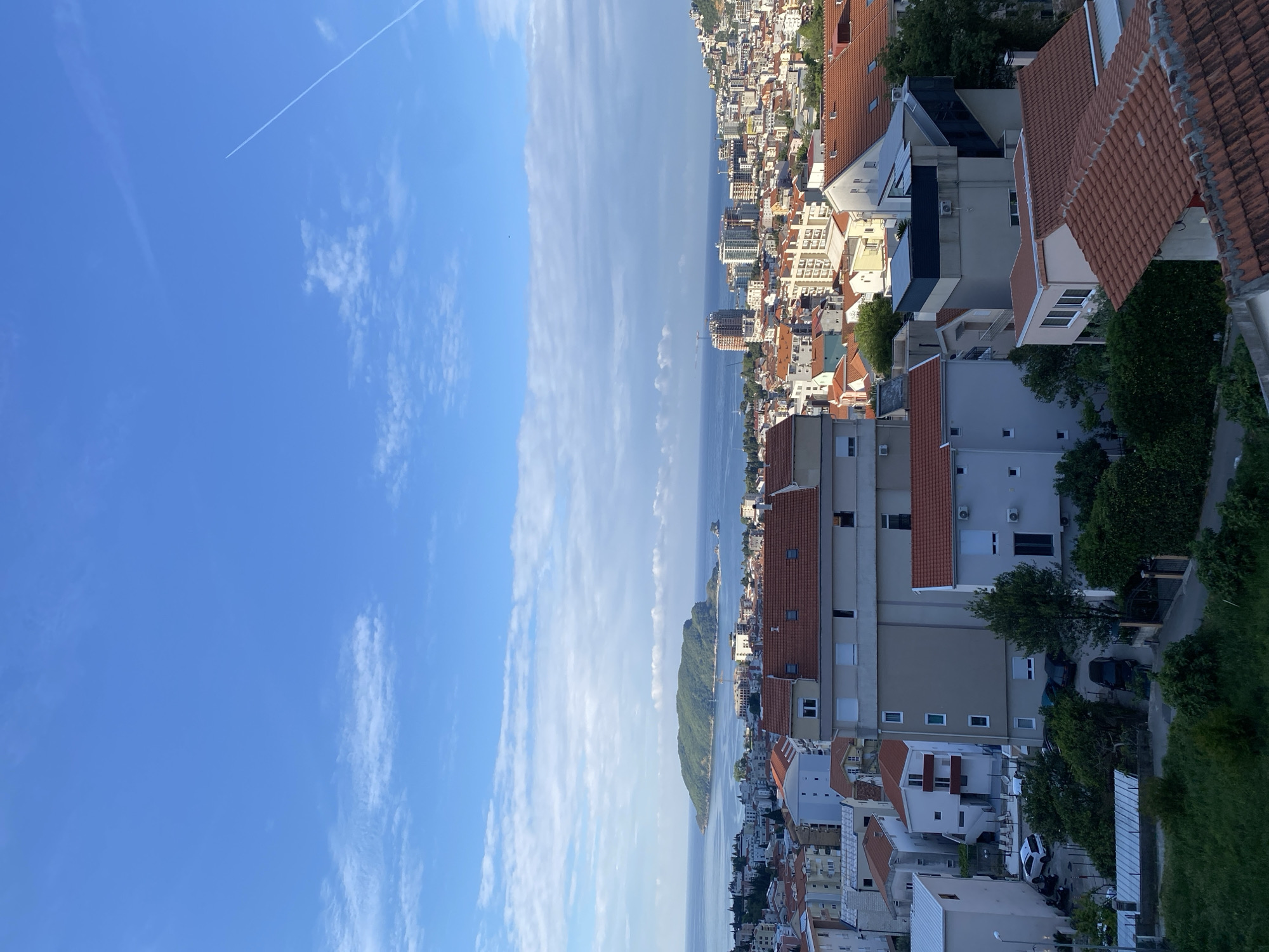 The image shows an aerial view of Budva, Montenegro, on a clear day. A blue sky with thin, wispy clouds and a contrail is visible. Below, a narrow strip of the Adriatic Sea separates the sky from the cityscape. Buildings with red-tiled roofs are prominent. A small, green, mountainous island sits in the water. The perspective suggests the photograph was taken from a high vantage point in the city. Cars and trees can be seen among the buildings and streets.