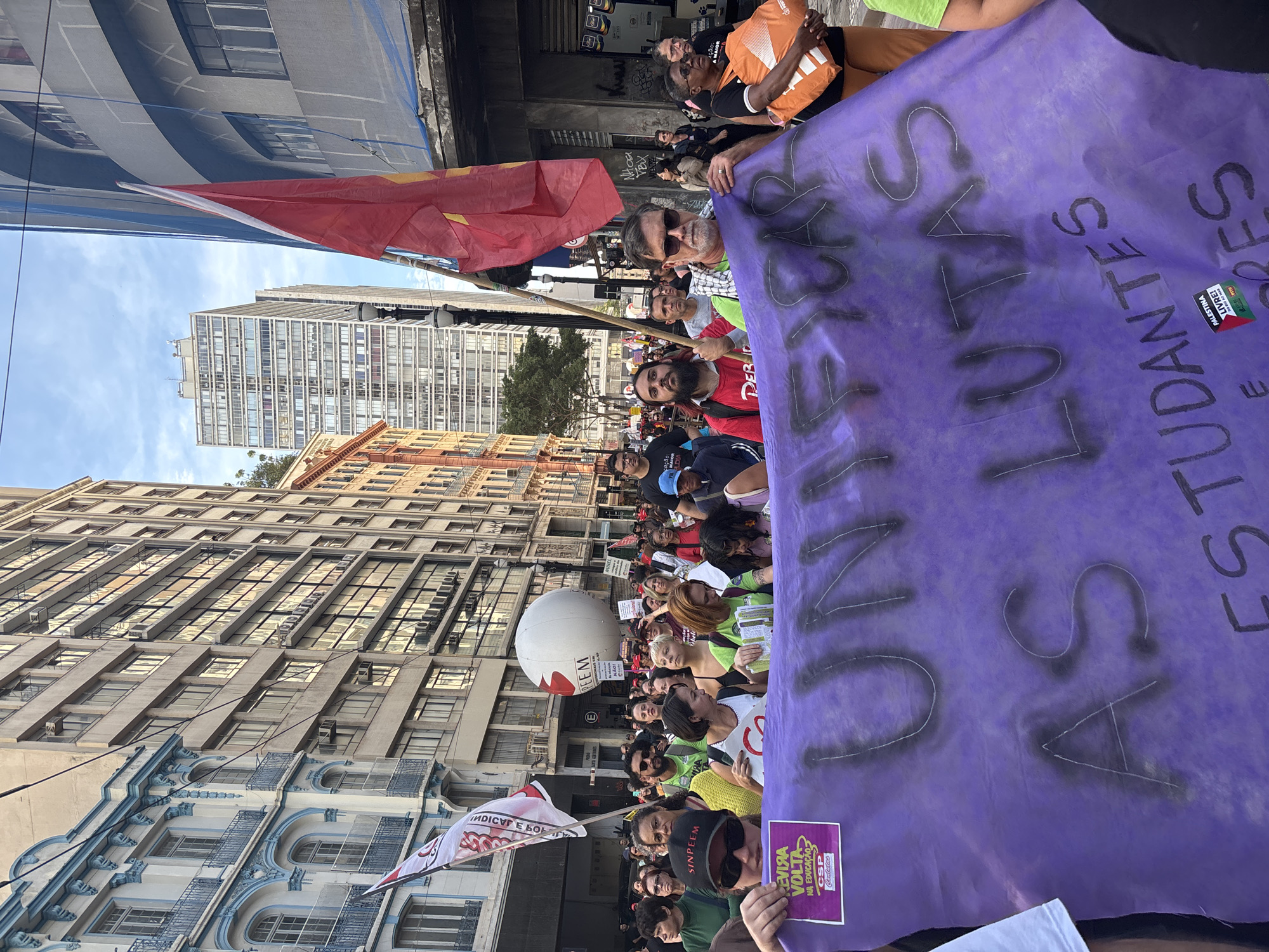 A medium-shot, eye-level captures a protest procession on a street flanked by tall buildings in São Paulo, Brazil. The crowd, varying in age, is tightly packed, holding aloft a large, vertical, purple banner with the message "UNI AS LUTAS ESTUDANTES". A red flag waves in the background to the left, partially obscuring the buildings. A white balloon with the words "PEEM" in red font is also visible in the center of the crowd. Some protestors carry additional signs. The architectural backdrop is a mix of old and modern buildings with ornate facades. The sky is partly cloudy.