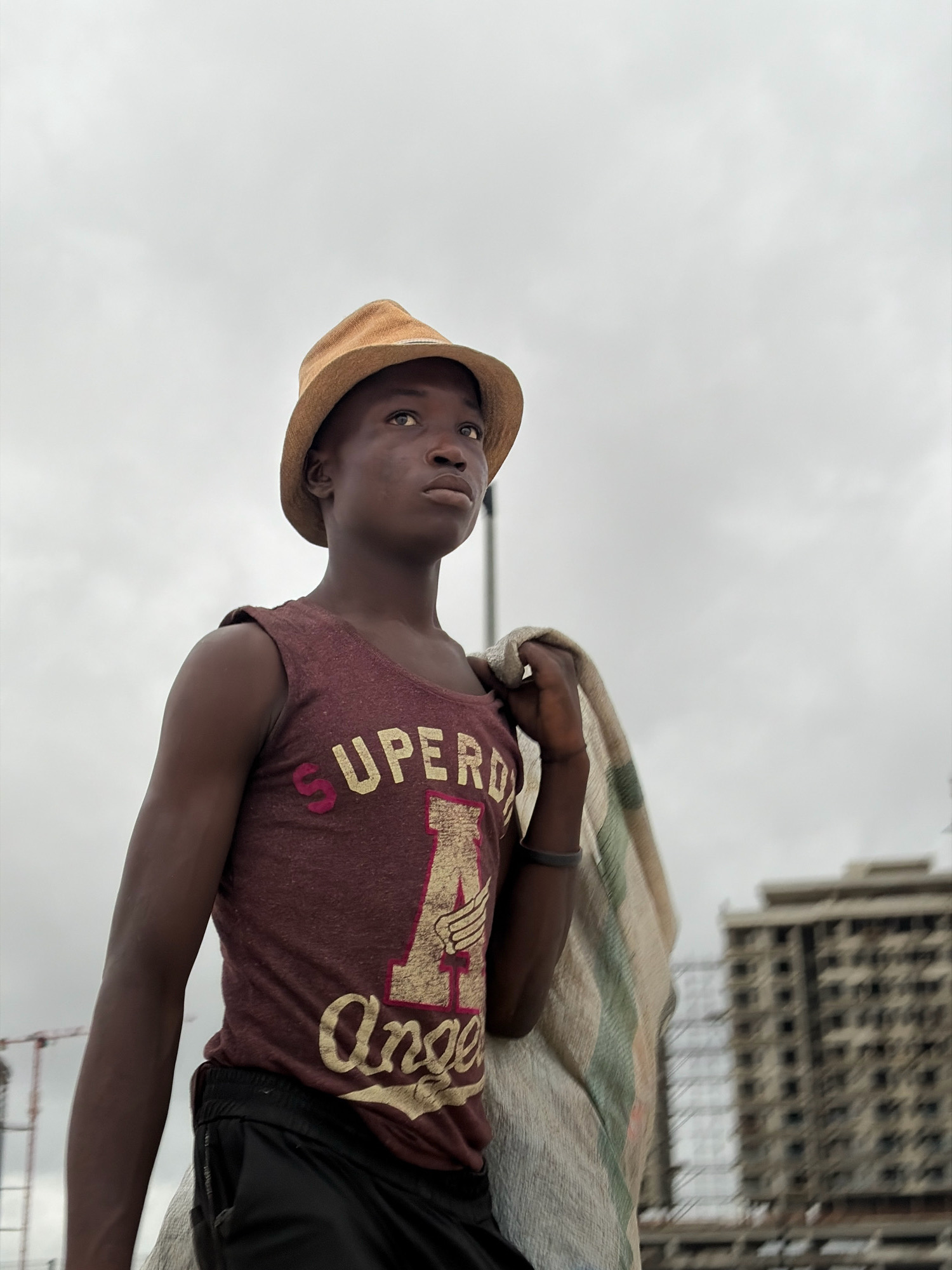 A low-angle, medium shot captures a young Black male from the chest up, wearing a tan fedora and a maroon tank top with "SUPERDRY" and an eagle emblem in yellow and pink. He carries a rolled-up striped blanket over his right shoulder. He is looking upwards and to his right, with a neutral expression. The background is a hazy, overcast sky, with the blurry outlines of a tall, unfinished concrete building and construction cranes on the right and left, respectively, suggesting an urban environment in Alaguntan, Nigeria.