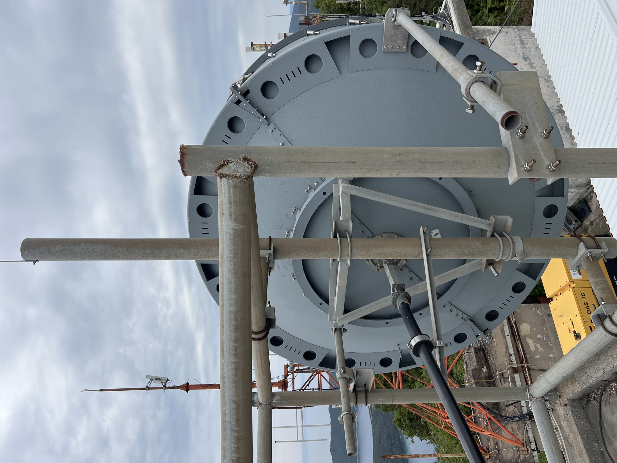 The image shows a large, gray, circular structure atop a building, likely part of a telecommunications installation. The structure is supported by a scaffolding-like framework of metal pipes. The location is indicated as Budva, Montenegro, based on context clues (though no specific landmarks are present in the photo).
The circular structure has a series of holes and slots around its perimeter. A thick black cable runs down from underneath the structure. Some yellow equipment boxes labeled "OAS 78" are visible to the right. An orange tower framework is partially visible in the background. The sky is cloudy. There are no people in the image.