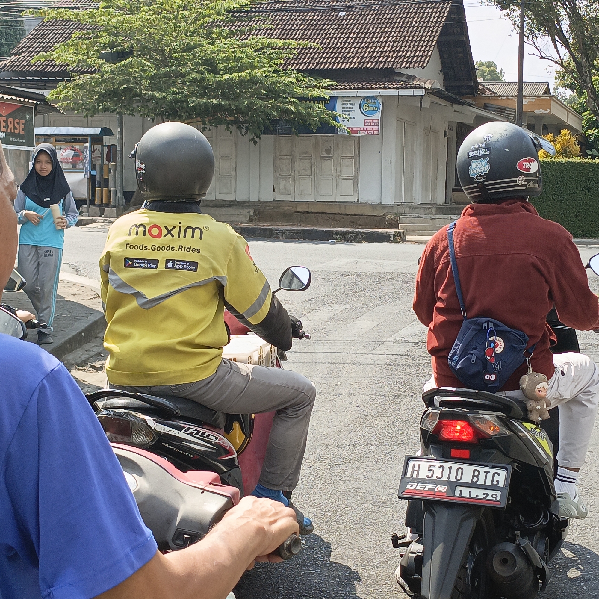 In Salatiga, Indonesia, two individuals are seen on motorcycles in traffic. The person in the foreground, wearing a blue shirt, has their left arm and hand visible, gripping a motorcycle's handlebar. Behind them, another individual in a yellow jacket with "Maxim Foods. Goods. Rides" emblazoned on it, is riding a motorcycle. This person wears a black helmet and grey pants. To the right, a third individual, wearing a maroon shirt and white pants, is also on a motorcycle, carrying a small blue bag with cartoonish charms. Their motorcycle has a license plate reading "H 5310 BTG" and "11.29". A young woman in a blue top and grey pants, wearing a hijab, stands on the sidewalk to the left, holding a rolled-up object. The background features buildings with tiled roofs, a tree with dense green foliage, and a street with asphalt pavement. The lighting suggests a sunny day.