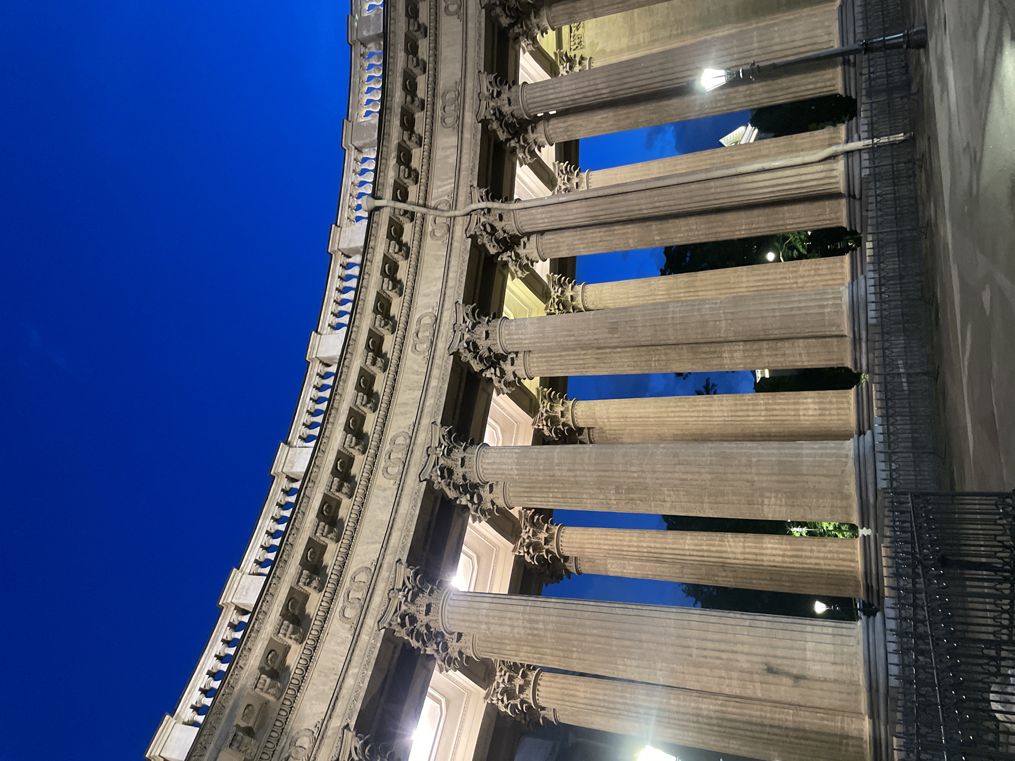 The image shows a low-angle, vertical view of the Kazan Cathedral in Saint Petersburg, Russia. The sky is a deep, clear blue, indicating twilight or night. The foreground features a dark metal fence. Behind the fence, a row of large, ornate Corinthian columns stretches upwards, supporting a curved, multi-layered architectural facade. The columns are illuminated, casting shadows and highlighting their fluted texture. Between some of the columns, glimpses of greenery and a distant building with a light can be seen. A vintage-style street lamp stands to the right of the frame, also illuminated. A thin, pipe-like structure extends vertically down the facade from above the columns. The overall scene conveys a sense of grandeur and historical architecture under a twilight sky.
