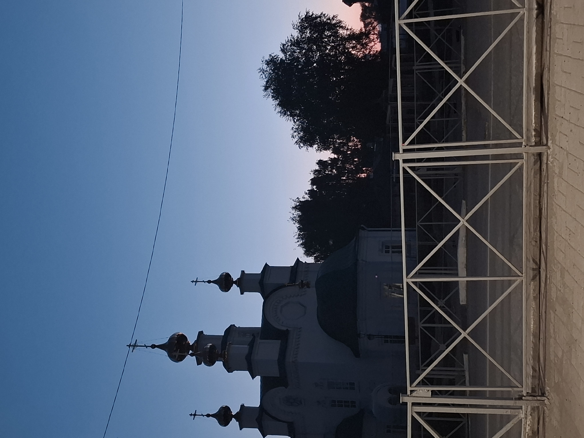 The image shows a white Orthodox church with golden domes and crosses against a twilight sky. A metal fence with a diamond pattern is in the foreground. Trees are visible behind the church. The scene appears to be in Kungur, Russia, likely near the Transfiguration Cathedral, which has a similar architectural style and golden domes. The lighting suggests it is either dawn or dusk. There are no people or animals visible in the image. The overall impression is one of quiet serenity and architectural beauty.