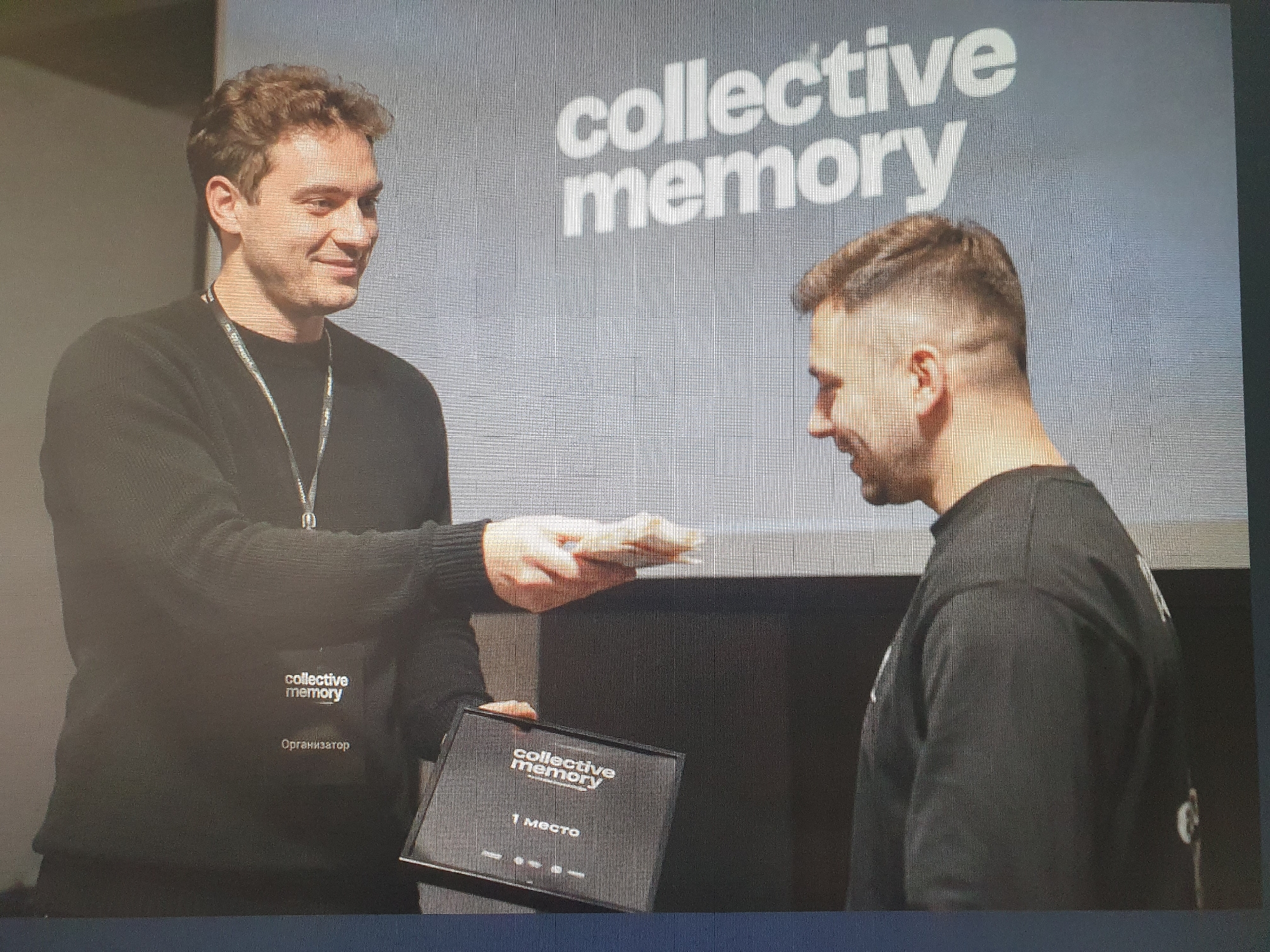 Two men are on a stage in front of a screen that displays the words "collective memory" in large white letters. The man on the left, wearing a black sweater with a lanyard and a badge that reads "collective memory" and "Организатор" (Organizer), is handing a black box with "collective memory" and "1 место" (1st place) written on it to the man on the right. The man on the right is wearing a black long-sleeved shirt and has a shaved head with a fade haircut. The man on the left is smiling and looking at the man on the right. The man on the right is looking down at the box. The environment appears to be an indoor event space. The image does not provide any information that would allow for the identification of a specific location in Tashkent, Uzbekistan.