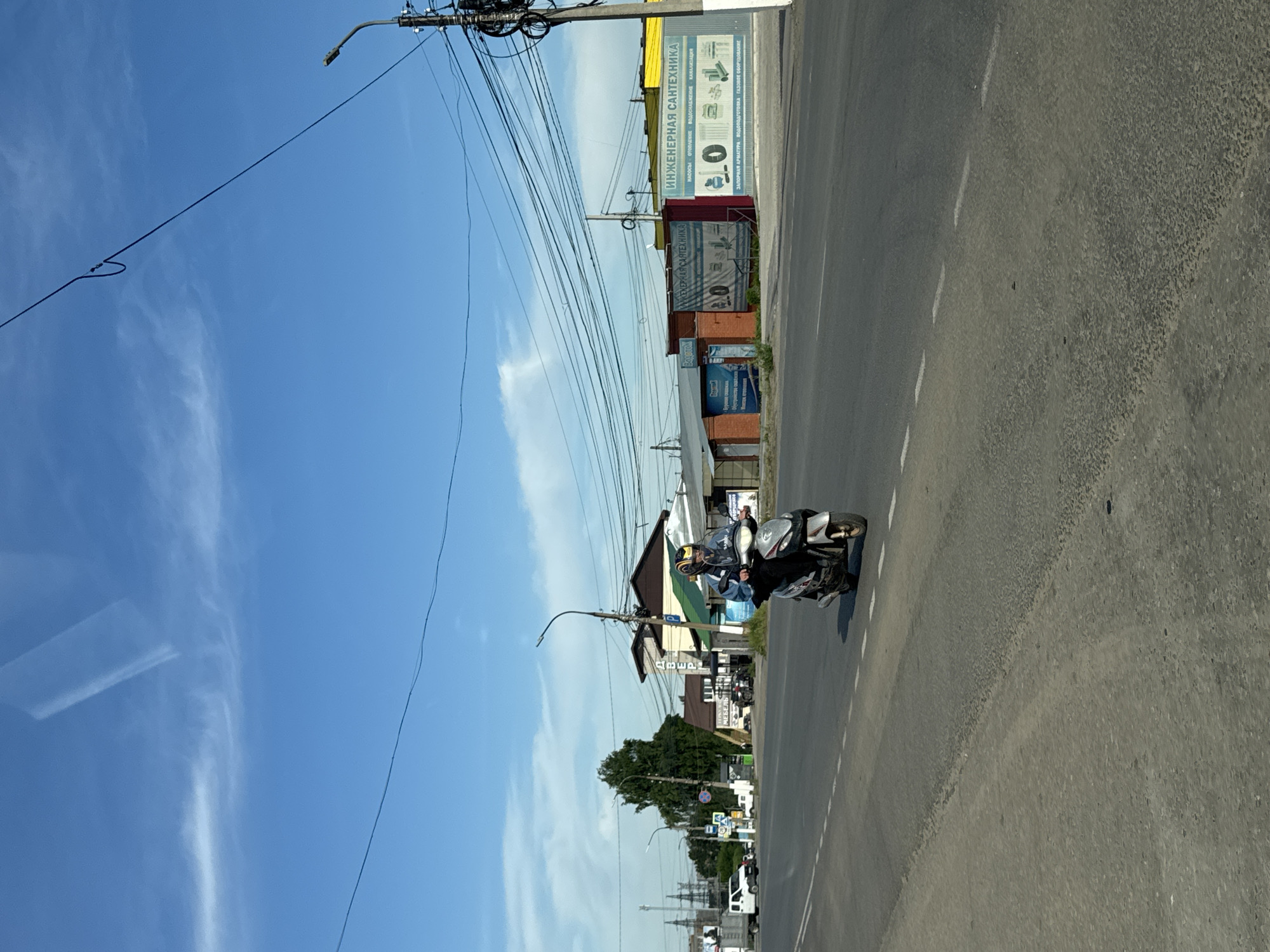 A person rides a scooter on a two-lane asphalt road under a bright blue sky with scattered white clouds. The scooter rider wears a helmet and a blue jacket. Several buildings are visible on the right side of the road, including a structure with a yellow facade and the words "ИНЖЕНЕРНАЯ САНТЕХНИКА" (ENGINEERING PLUMBING) in large blue letters. To the left of the road, there is a white truck, a large green tree, and more buildings. A utility pole with numerous wires is on the right side of the image. The scene appears to be a street in a developed area, likely a town or city. The presence of overhead power lines and commercial buildings suggests a functional urban environment.