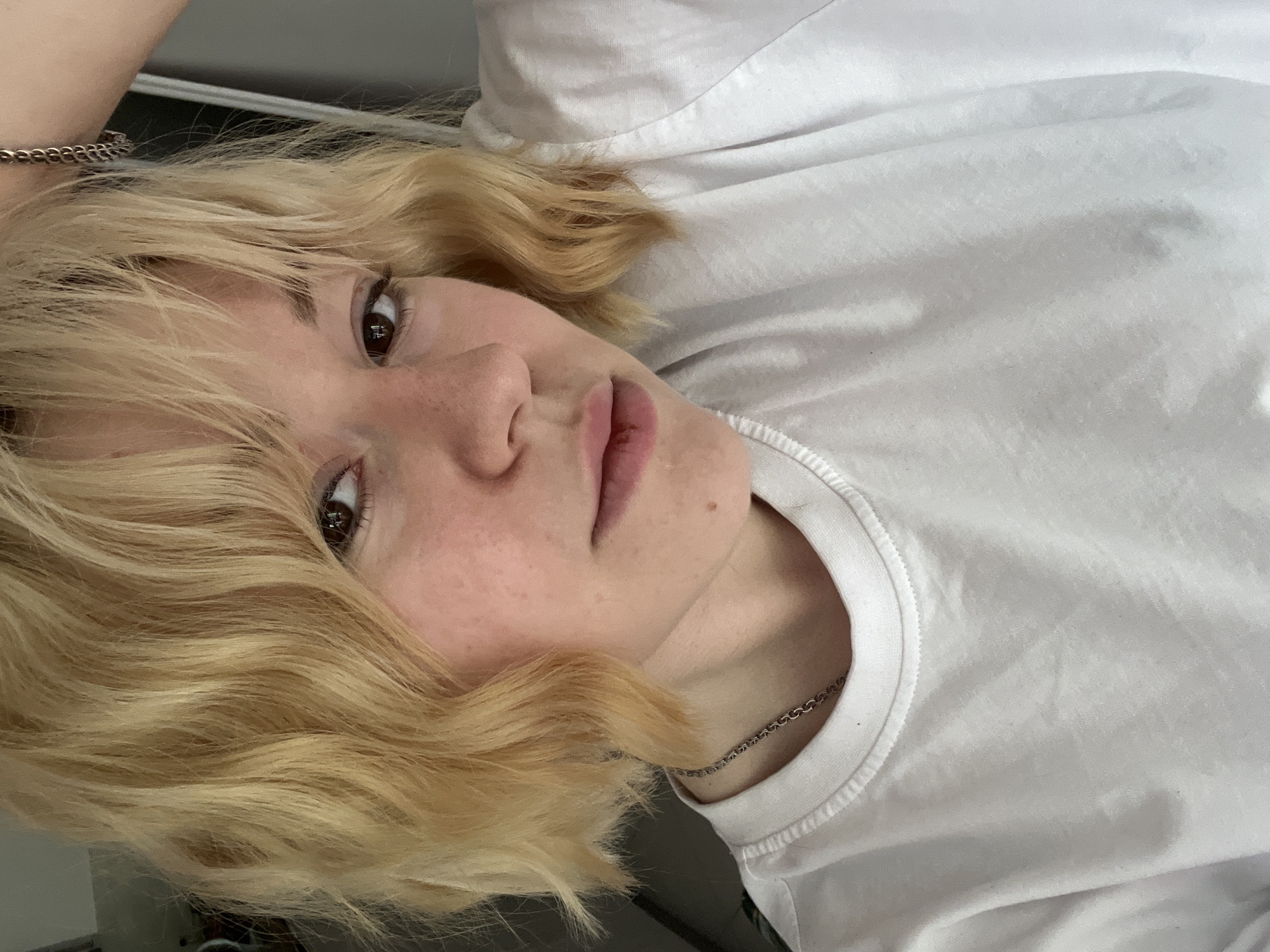 A person with light blonde hair is pictured from the chest up. The person is wearing a plain white t-shirt and a thin silver chain around their neck. They have brown eyes and fair skin. The background is out of focus. The location is unknown, and there is no visible indication that the image was taken in Boreti, Montenegro.