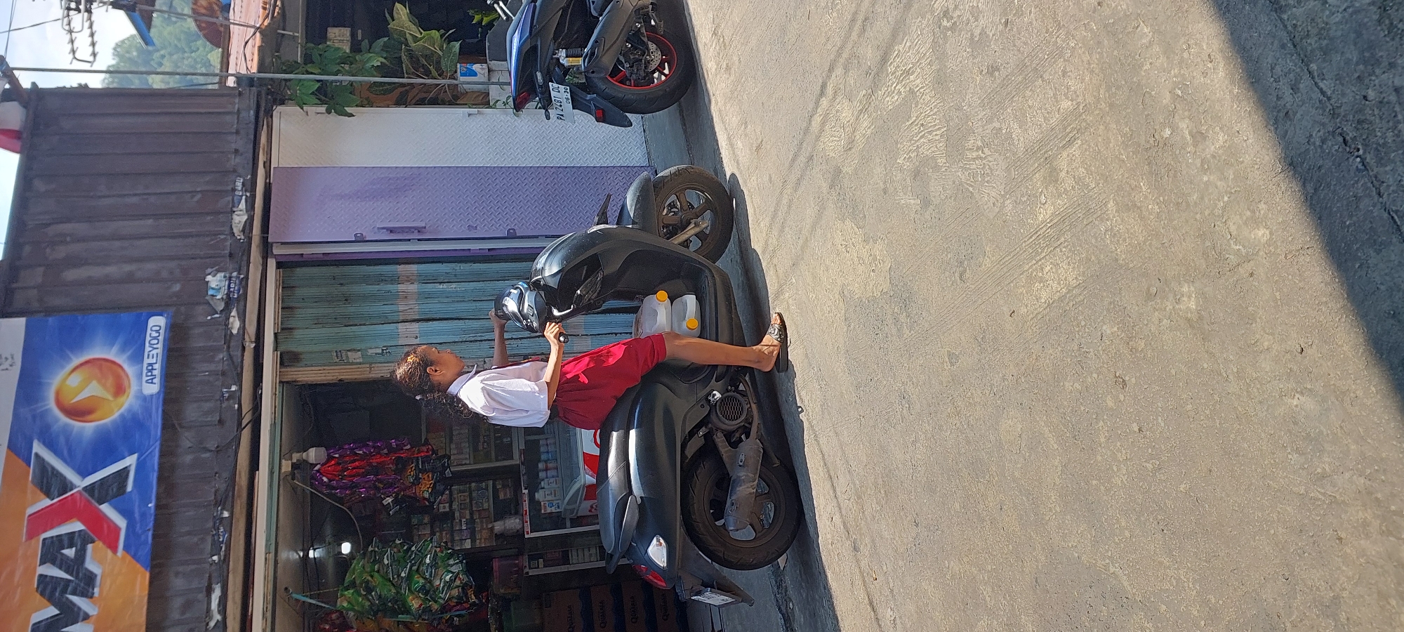 An Indonesian schoolgirl in a white shirt and red shorts is sitting on a black scooter outside a shop in Jayapura, Indonesia. The shop has a sign that reads "MAX APPLE YOGO" and appears to be a small convenience store. The girl is holding the scooter's handlebars. Another scooter is parked behind her to the right. The ground is a paved road with some tire marks. The background shows a building with a corrugated metal roof and some vegetation.