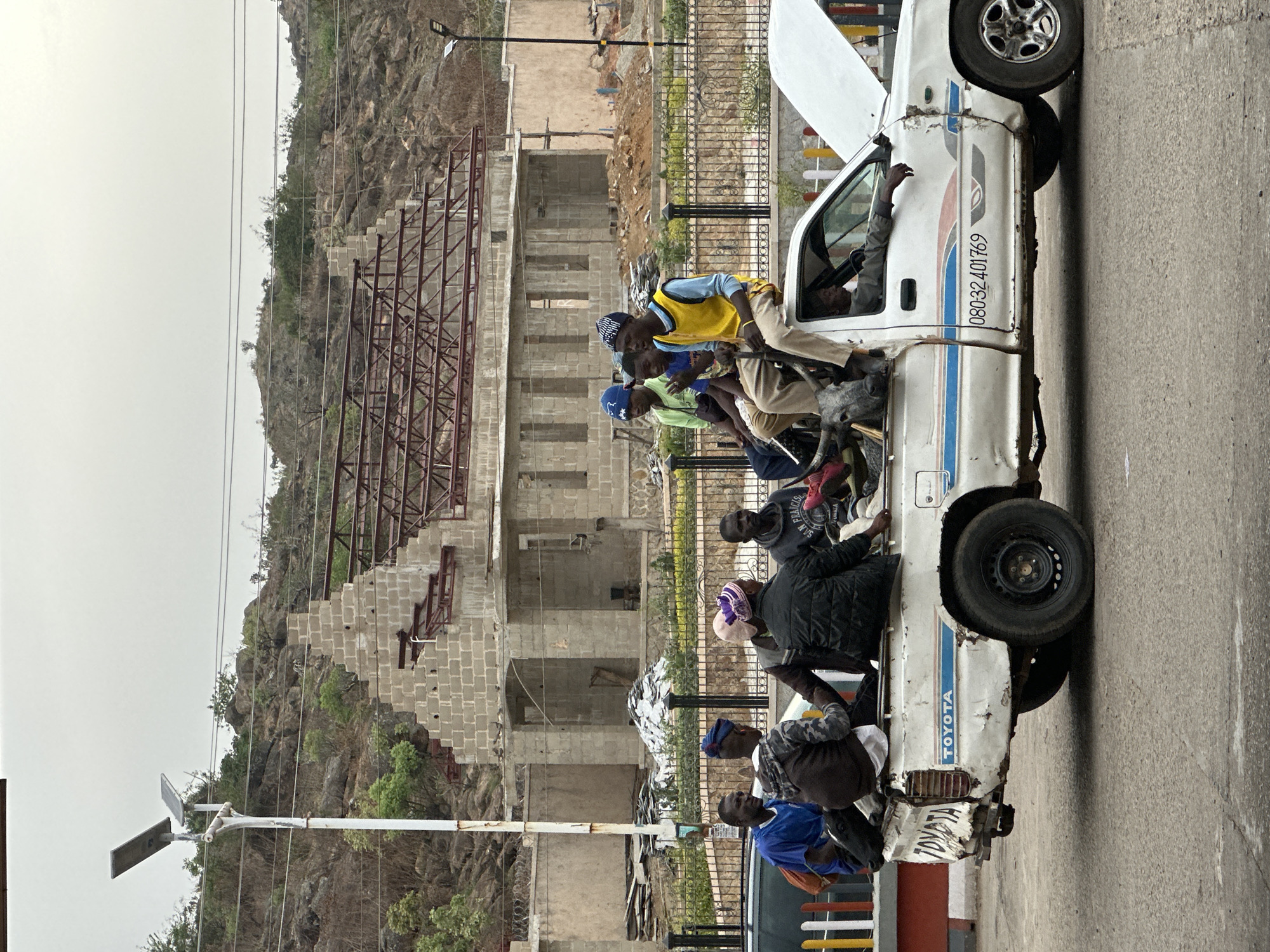 The image captures a lively street scene, likely in Jos, Nigeria, featuring a heavily loaded white Toyota utility vehicle. The vehicle, possibly a pick-up truck or a shared taxi, shows significant wear and tear, with dents and an open hood, suggesting it is either stopped for maintenance or waiting for passengers. Several men, numbering at least ten, are visible on and around the vehicle, indicating an overloaded state characteristic of informal public transport. Some passengers sit precariously on the side rails of the truck bed, while others are inside the cab; one man with a yellow vest is prominent towards the front, and the driver's arm is seen extending from the cab window.
The side of the vehicle prominently displays the word "TOYOTA" in vertical lettering, along with a contact number: "08032401769". A blue and red stripe also runs along the vehicle's body.
In the background, a rugged, rocky hill rises, partially covered with sparse, dry vegetation. At the base of the hill, an unfinished concrete block building with exposed rebar structures signifies ongoing construction. A metal fence separates the road from this area. The sky is overcast or hazy, providing soft, diffused lighting to the scene. Utility poles carrying power lines and a street light with attached solar panels are also visible, hinting at the infrastructure of the area. The overall scene depicts a functional, everyday moment of travel and development in the region.