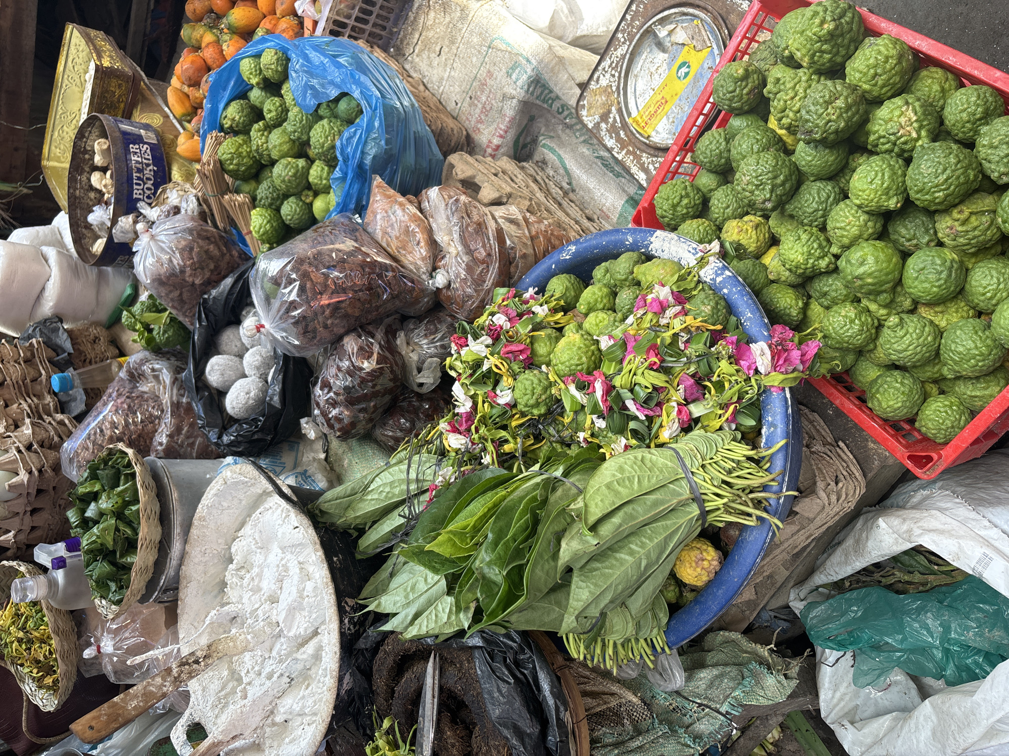 This is a high-angle, eye-level shot of a market stall in Bireuen, Indonesia, displaying various agricultural and food products. In the foreground, a red plastic crate is overflowing with kaffir limes, their bumpy green skin and irregular shapes clearly visible. To the left of the crate, a blue bowl contains more kaffir limes mixed with colorful flower petals and green leaves, some of which appear to be betel leaves. Behind the blue bowl, a large pile of bundles of green leaves, likely betel leaves, are tied with black elastic bands, with their stems pointing downwards.
Further back, a large metal pan is filled with white flour or a similar powdery substance, with two wooden utensils submerged in it. To the left of the pan, a black plastic bag contains dark, dried material. Scattered around are numerous plastic bags filled with various dried goods, appearing as brown, granular, or fibrous textures. Several woven baskets are visible, one containing yellow, leafy material, and another holding small, green, pyramid-shaped items wrapped in leaves. In the background, a blue can with the label "BUTTER COOKIES" is visible, along with other food containers and bags. The overall environment suggests a bustling outdoor market setting with an abundance of fresh produce and packaged goods.