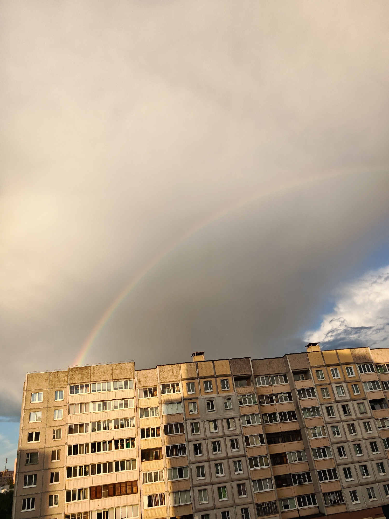 A wide shot captures a residential building in Minsk, Belarus, under a dramatic sky with a rainbow. The building is a multi-story, concrete structure with rows of identical windows. The sky is a mix of pale yellow clouds, darker gray storm clouds, and a sliver of blue sky with white cumulus clouds on the right. A visible rainbow arcs across the upper portion of the frame, partially obscured by the clouds. The building's facade shows signs of wear, with some areas appearing faded or discolored. The windows are mostly closed, with some reflecting the light from the sky. The scene suggests a post-rain or approaching storm weather event.