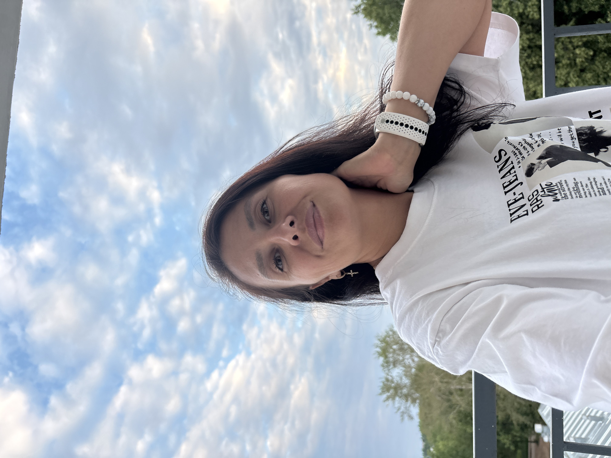 A woman is taking a selfie outdoors, with a cloudy sky in the background and trees visible. She is wearing a white t-shirt with text and graphics printed on it. She has dark hair and is wearing bracelets on her left wrist and a small cross earring. The image is oriented vertically, suggesting it may have been taken with a phone. The visible text on her shirt includes "EVE-JEANS" and "RAS," along with other fragmented words and numbers that appear to be part of a design. The scene suggests a casual, daytime setting, possibly in a park or on a balcony, given the presence of trees and railings. The lighting suggests it is daytime, with the sun possibly behind the clouds.
