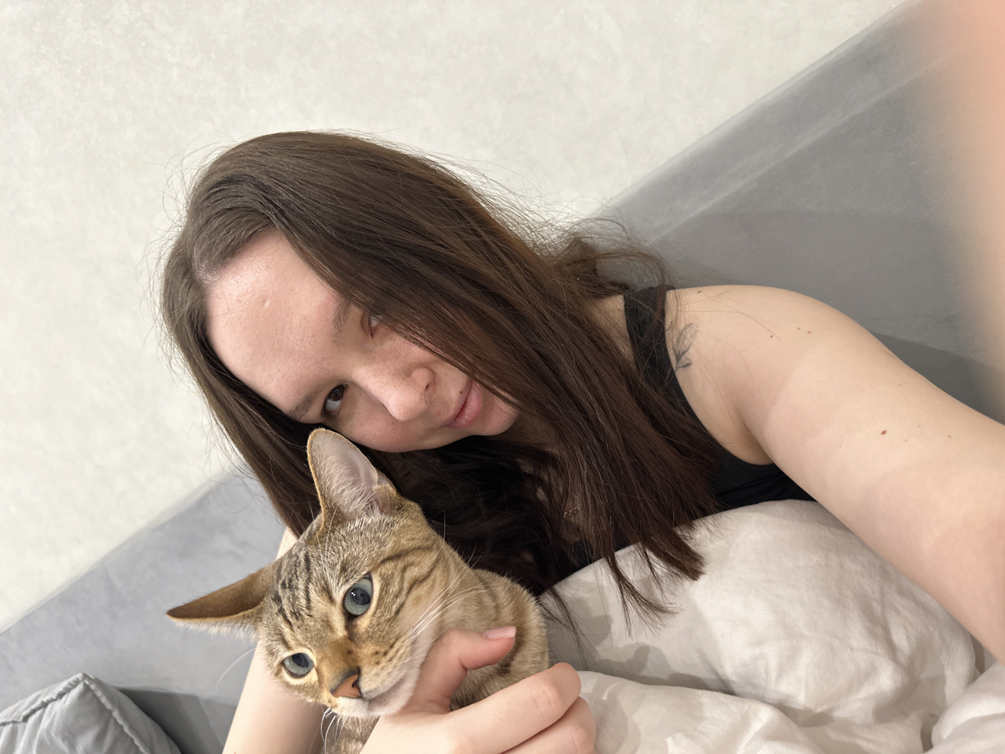 The image displays a close-up indoor scene featuring an adult female embracing a tabby cat. The female, visible from the chest up, has long, dark brown hair covering part of her face, with her left eye, freckled nose, and mouth visible, exhibiting a slight smile. Her left arm supports the cat, with her hand gently around its neck. A faint tattoo is present on her left upper arm. She wears a dark-colored garment. The cat, with short, striped brown fur and light blue-green eyes, is looking directly at the camera, held close to the female's body. Both subjects are positioned against a white, wrinkled duvet or blanket that covers the lower left of the frame. The background consists of a light-colored wall with a subtle texture and a portion of a grey surface, possibly a pillow or headboard, in the upper left and lower right. The specific location within the city of Chaikovsky, Russia, is not discernible from the visual information in the image.