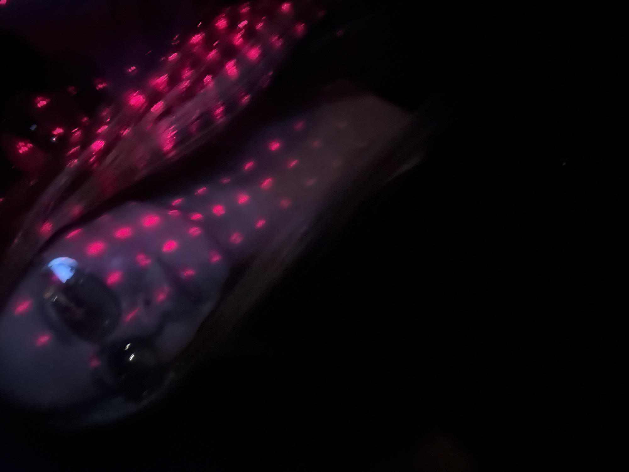 The image shows a person's face illuminated by pink dotted lights, possibly from a projector or a light show. The person is wearing glasses, and their eyes are visible through the lenses, reflecting some of the lights. The background is dark, with some blurry pink lights extending to the right of the person's head. The perspective is a close-up, angled shot. There are no discernible elements that would indicate a specific location in Chaikovsky, Russia.
