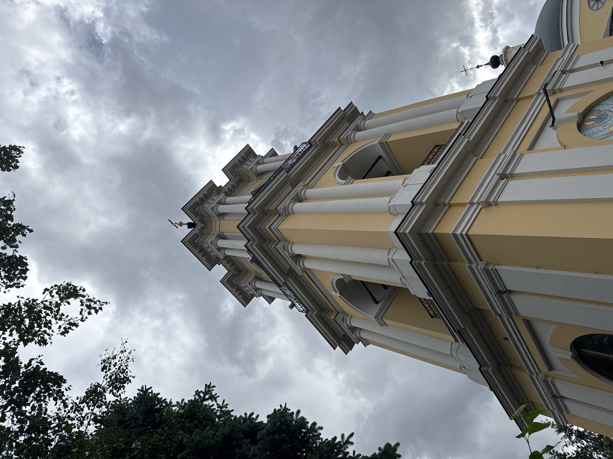 The image shows a low-angle view of a tall, multi-tiered bell tower or building facade with classical architectural elements such as columns, arches, and decorative moldings. The color scheme is predominantly yellow with white accents. At the top of the tower is a small dome topped with a cross. The sky behind the tower is overcast with gray clouds. In the lower left and bottom of the frame, there are trees. Based on the architectural style and details, the building is located in Moscow, Russia.