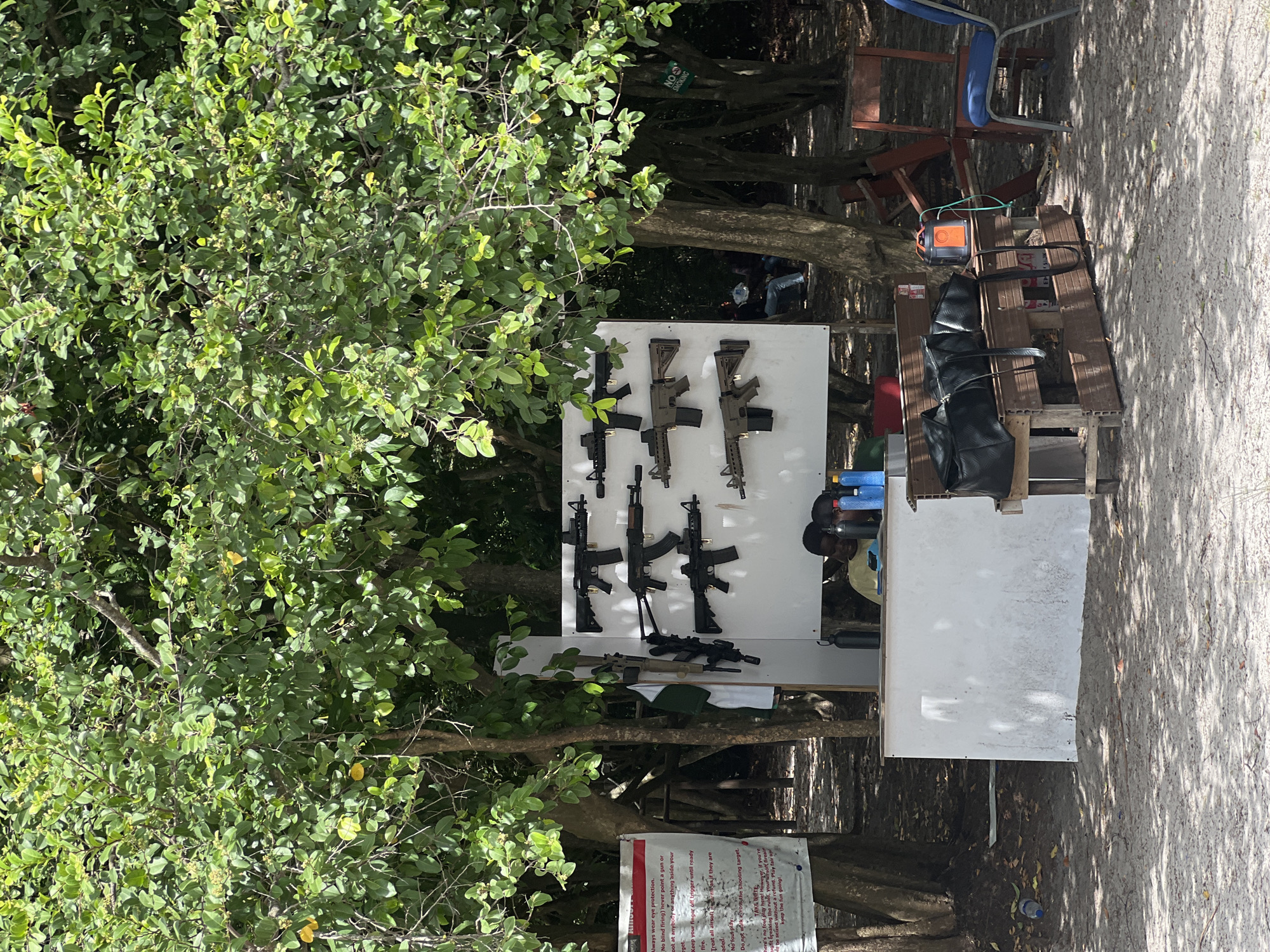 The image shows an outdoor airsoft shooting range in Lekki, Nigeria. A white display board features an array of airsoft rifles. A man is positioned behind a counter. Chairs and other equipment are nearby. Signage with safety rules is visible. The area is partly shaded by trees.