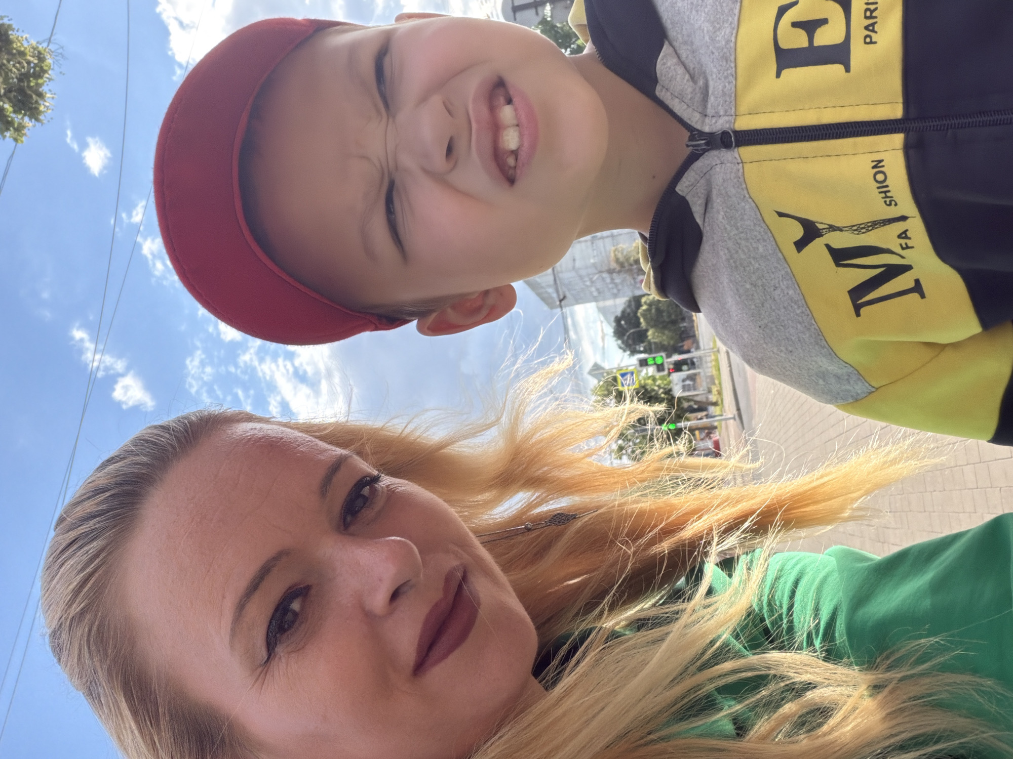 A selfie taken outdoors, possibly in Chernihiv, Ukraine, shows a woman and a young boy. The woman, on the left, has blonde hair and is wearing a green top. She is looking towards the camera with her head tilted. The boy, on the right, is wearing a red baseball cap and a grey and yellow hooded jacket with the text "MY FASHION PARIS" visible. He is making a grimacing face towards the camera, with his eyebrows furrowed and his teeth exposed. The background suggests an urban environment with a sidewalk, a street with traffic lights, and buildings. The sky is blue with some clouds, and there are power lines visible.
