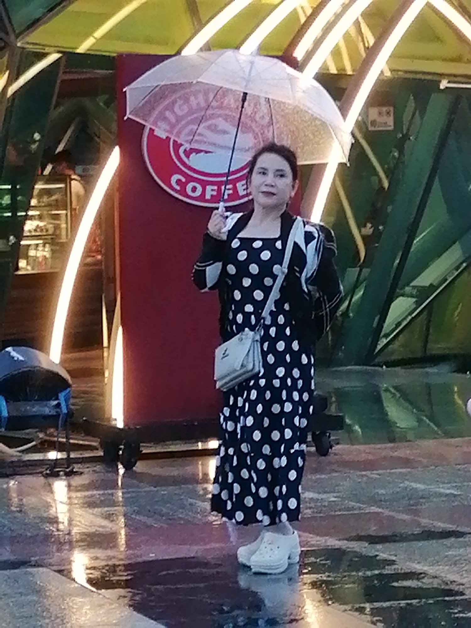 A woman stands in front of a red and white circular sign that reads "COFFEE" and what appears to be "HIGH" above it, possibly indicating a coffee shop in Da Lat, Vietnam. She is holding a clear umbrella, and wearing a polka-dot dress, a black and white jacket, and white clogs. The ground is wet and reflective, suggesting it has recently rained. There are also some pieces of outdoor furniture to the left of the woman and some green structural elements in the background.