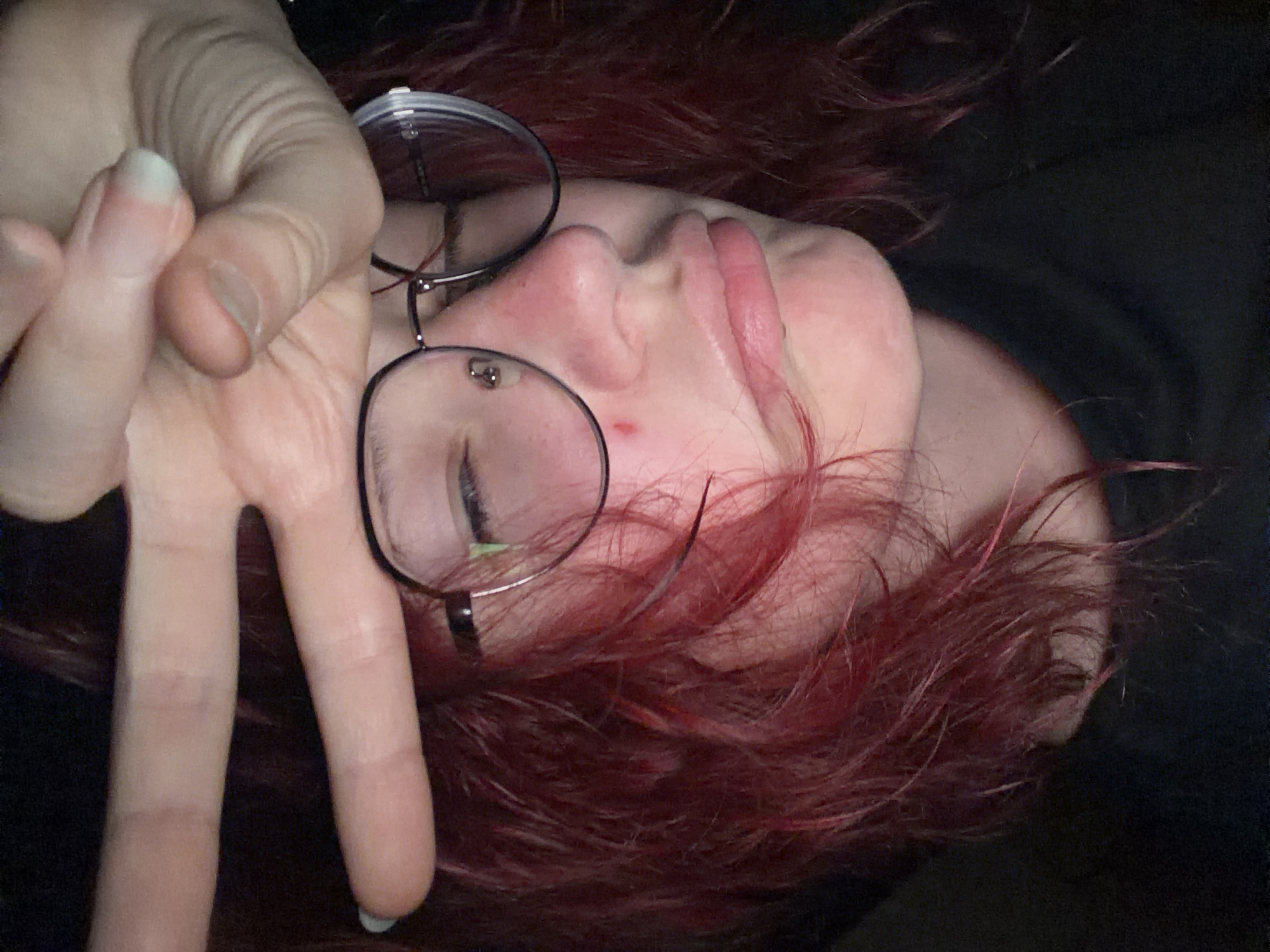 A close-up, low-angle, indoor, dimly lit, digital photograph shows a person with reddish-pink hair and dark-rimmed glasses. The person's eyes are closed, and their mouth is slightly open in a pout. A hand with painted nails is positioned over the person's forehead, with two fingers extended in a peace sign. The person appears to have a small red mark or blemish on their cheek. The background is dark and indistinct. No specific details about the city of Chelyabinsk, Russia are visible.