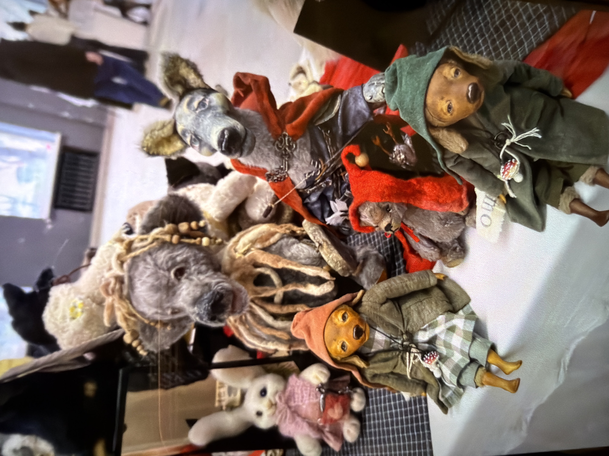 The image displays a collection of handcrafted dolls or stuffed animals arranged on a surface, possibly for sale or display. Several of the figures appear to be dressed in elaborate, vintage-inspired clothing and accessories. Some of the dolls have animal features, such as wolf or dog-like heads, while others seem to represent human-like characters. The scene includes a rabbit doll, and a few of the figures are adorned with beads, hoods, and other decorative elements. The presence of these handcrafted items suggests the scene could be a craft fair, market, or a specialized shop in a tourist-oriented area in Moscow, Russia. Without additional visual references, providing a pinpoint location within Moscow is not possible.