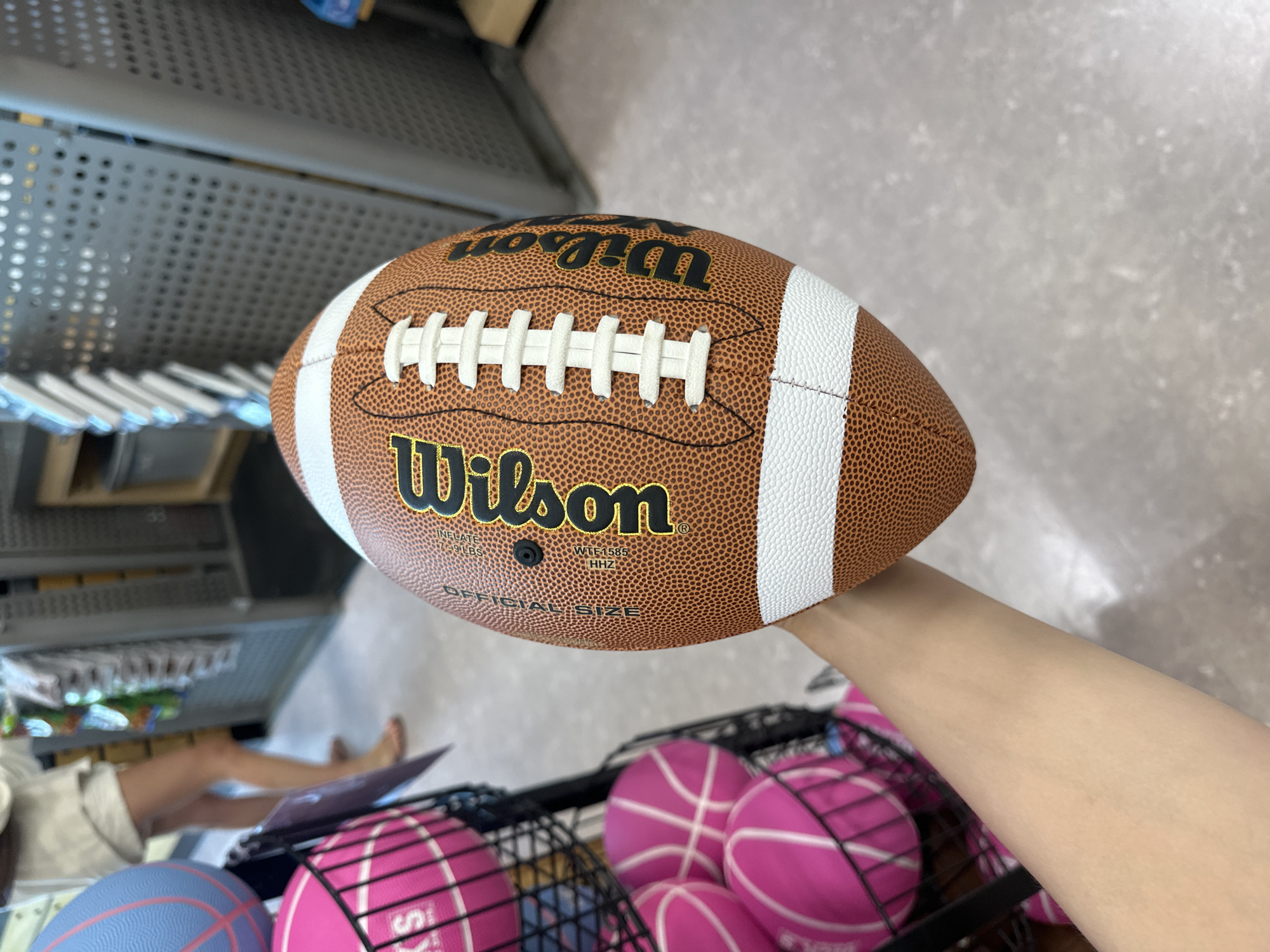 An over-the-shoulder medium shot captures a person's forearm holding a brown and white Wilson football. The football is the main subject, positioned in the center of the frame, with its textured surface and signature laces prominently displayed. To the left of the football, a black wire basket is filled with several pink basketballs. In the background, a person wearing light-colored shorts and a white top is partially visible, standing in what appears to be a retail store. Racks with merchandise are also visible in the blurred background, suggesting a sporting goods store. The floor is a neutral grey, and the lighting is typical of an indoor commercial setting. The location is not explicitly mentioned within the image, but a general understanding of sporting goods stores and their common placement within retail areas is implied.