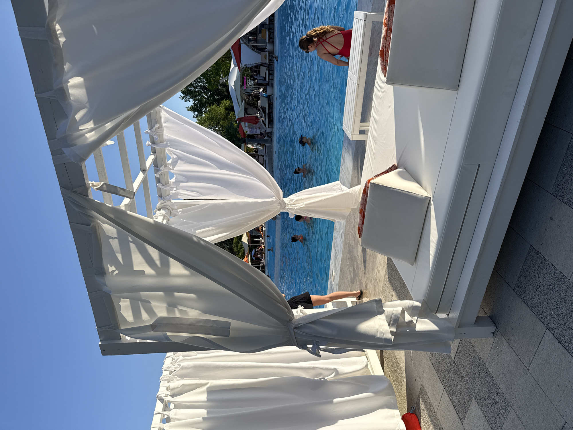 The image depicts an outdoor leisure area at a resort in Rostov-on-Don, Russia. In the foreground, a white cabana with flowing white curtains partially obscures the view. Beyond the cabana, a pool with several people swimming is visible. A woman in a red swimsuit stands at the edge of the pool near a white daybed with white cushions and red-patterned accents. Other lounge chairs and umbrellas are also visible in the background. The sky is clear and blue. The scene suggests a sunny day at a resort.
